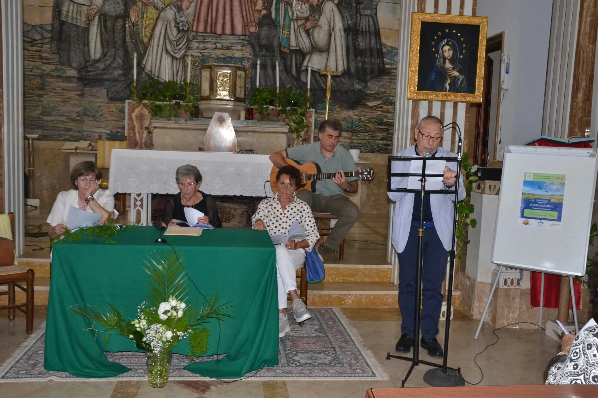 Un gruppo di persone sedute e in piedi in una chiesa, alcuni leggendo dalleggendo e altri ascoltando, con una persona che suona la chitarra e un'altra che canta davanti a un microfono. Sullo sfondo, un altare e un grande affresco religioso.