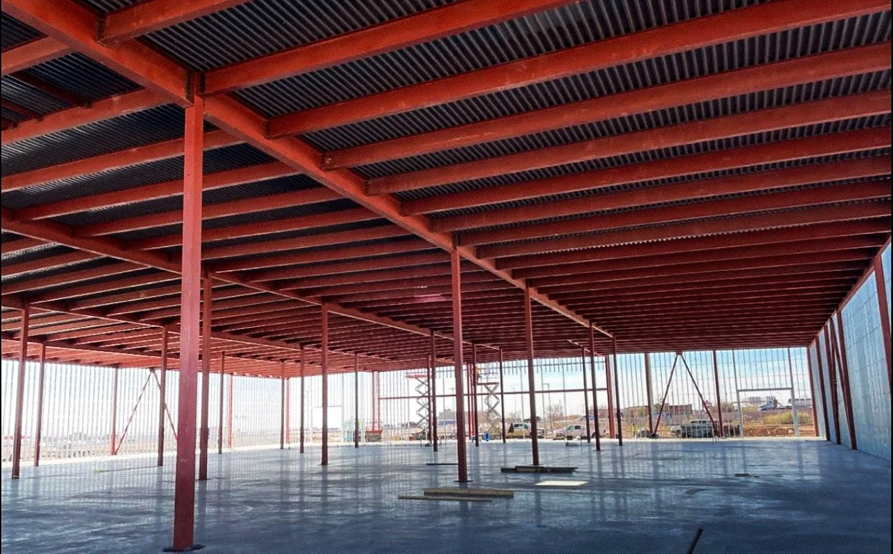 Construction site with a steel framework with red beams and an open floor, large windows in the background, and a view of a clear sky.