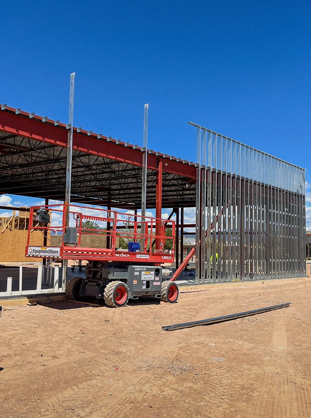 Construction site with a steel structure being assembled, a red scissor lift in the foreground, and a clear blue sky.