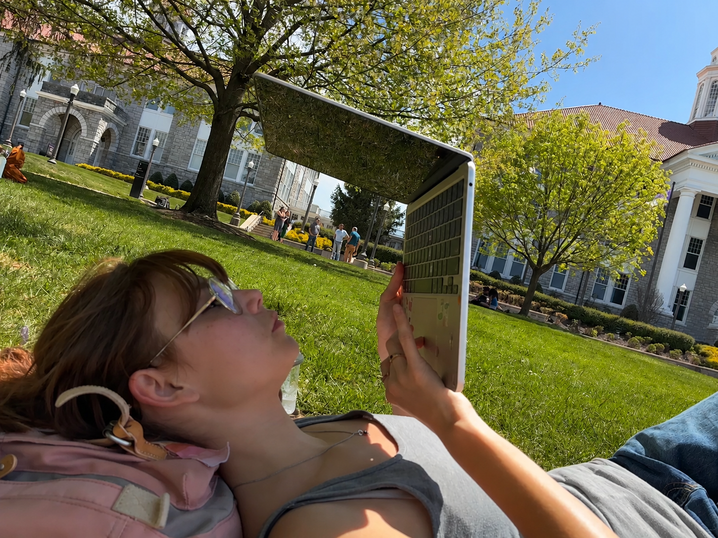 A young woman with sunglasses and a backpack lying on the grass outdoors, looking at her laptop computer, with a building, trees, and people in the background.