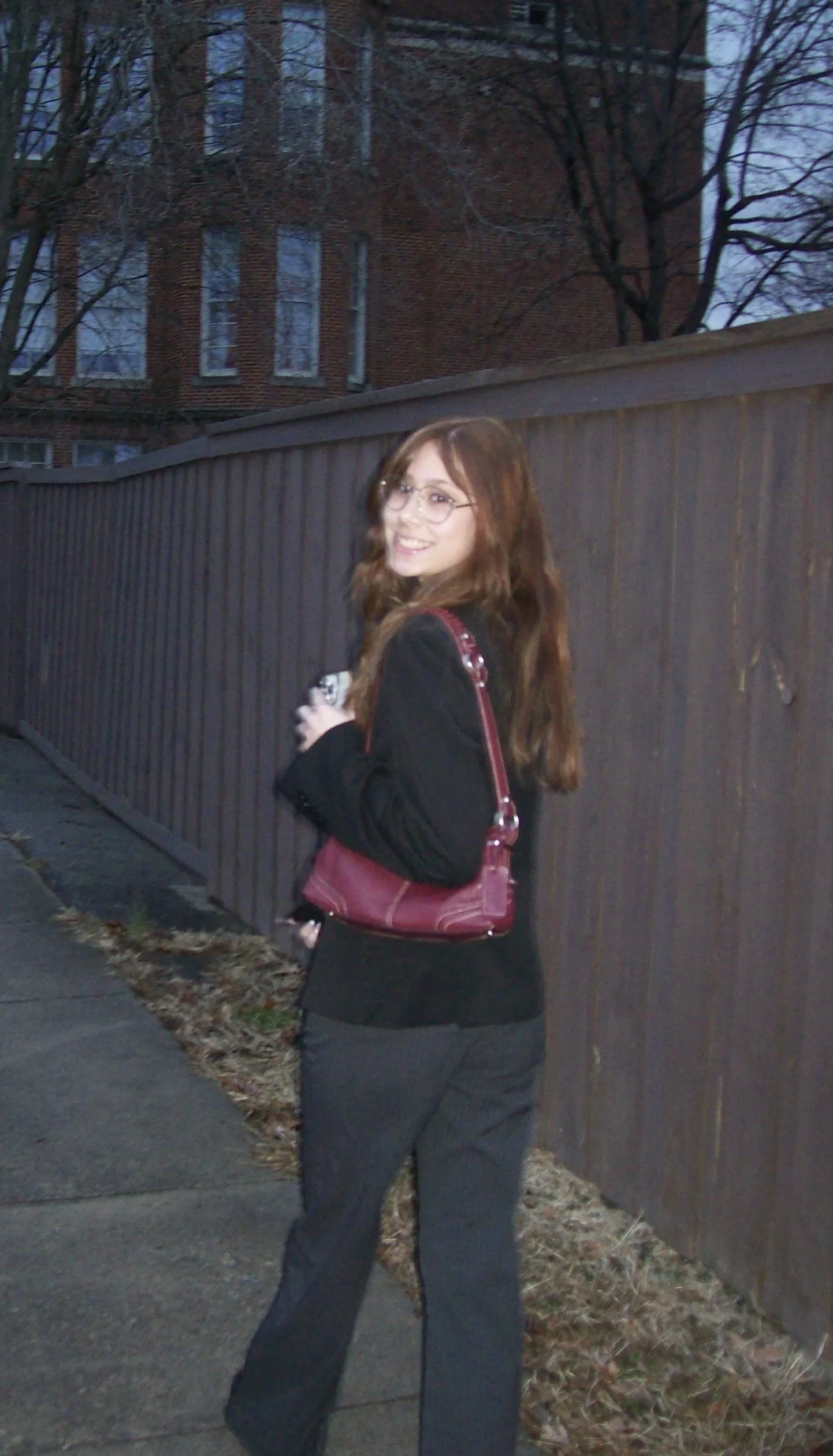 A woman with long brown hair, glasses, wearing a black jacket, gray pants, and carrying a red purse, stands on a sidewalk near a brown wooden fence, smiling at the camera with a multi-story brick building and leafless trees in the background during dusk.