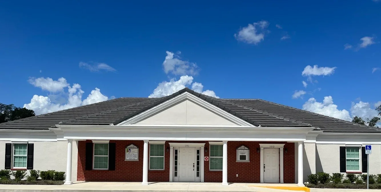 Root Cause FFM. A single-story building with white walls and red brick accents, supported by white columns, with a grey tiled roof, and a parking spot with a handicap sign in front.