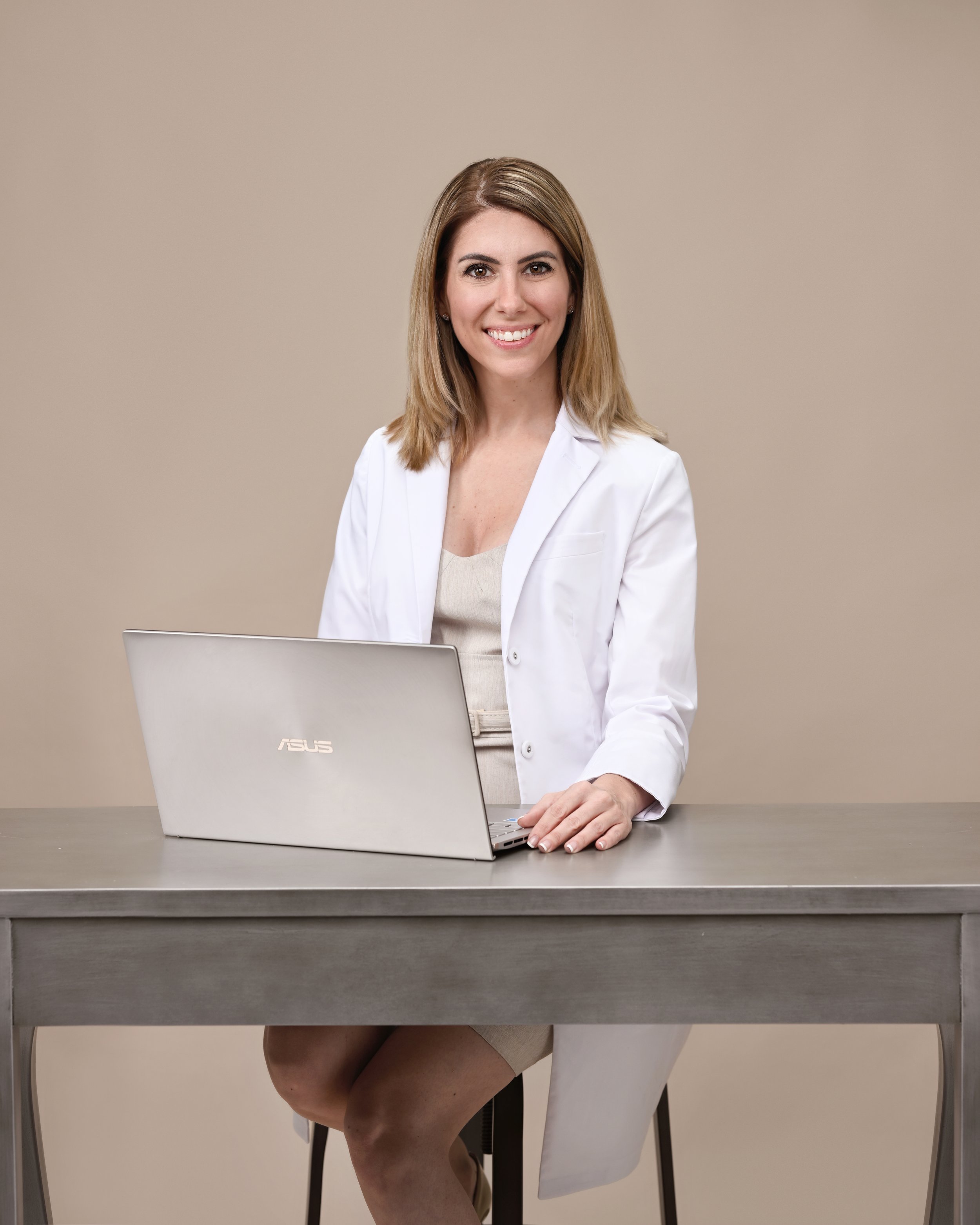 Dr. Turner, DNP sitting at a desk with a laptop, wearing a white blazer and beige dress, smiling at the camera.