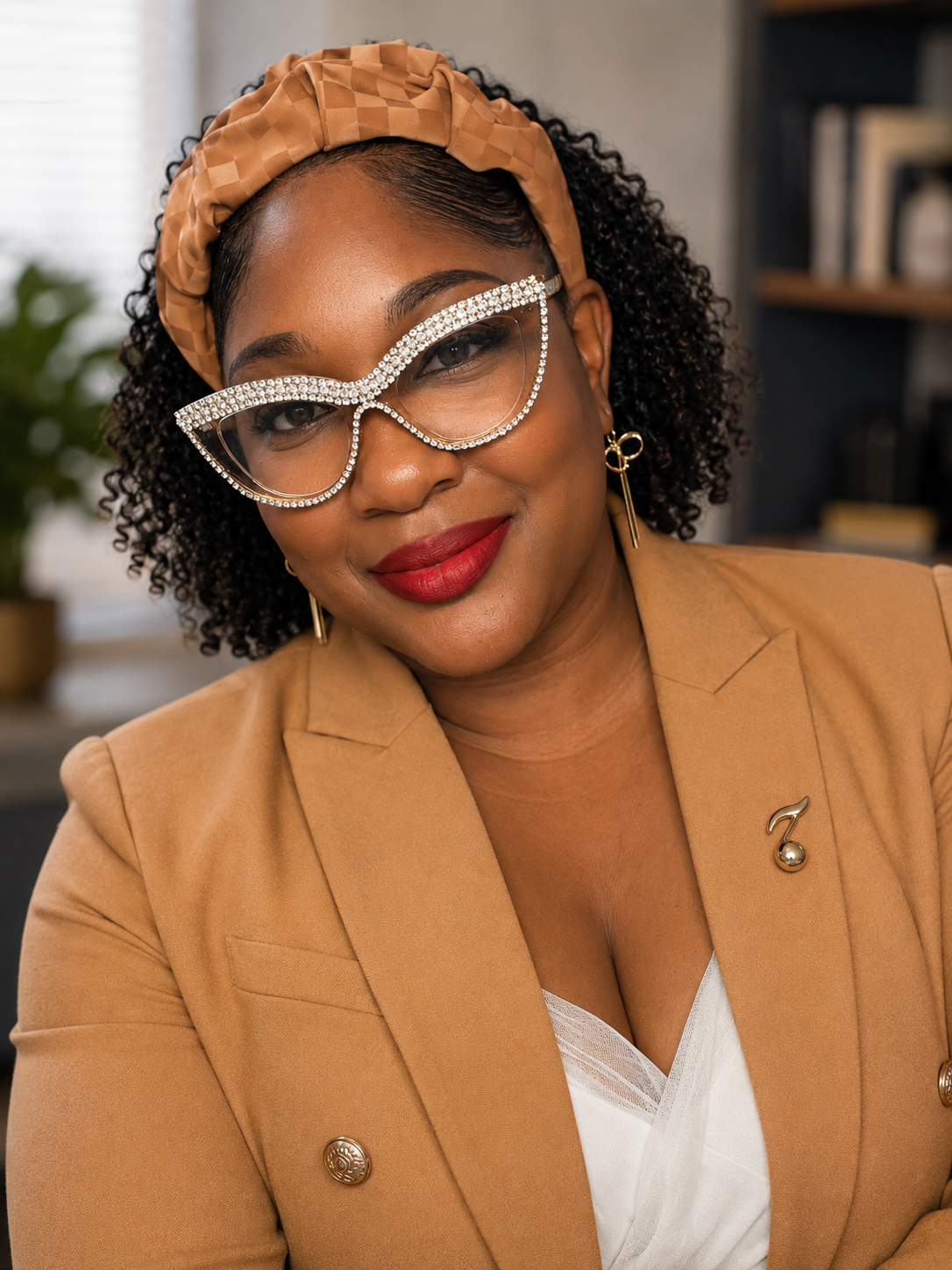 A woman with curly hair and glasses decorated with rhinestones, wearing a tan blazer with gold buttons and a brooch, and red lipstick, smiling in an indoor setting.