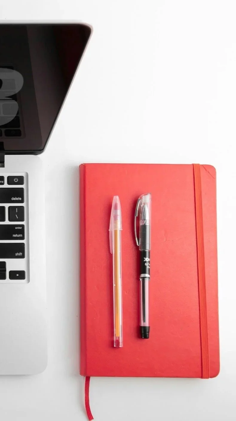 A white desk with part of a laptop on the left, and a red notebook with a red elastic band on the right, topped with a pink pen and a black pen.