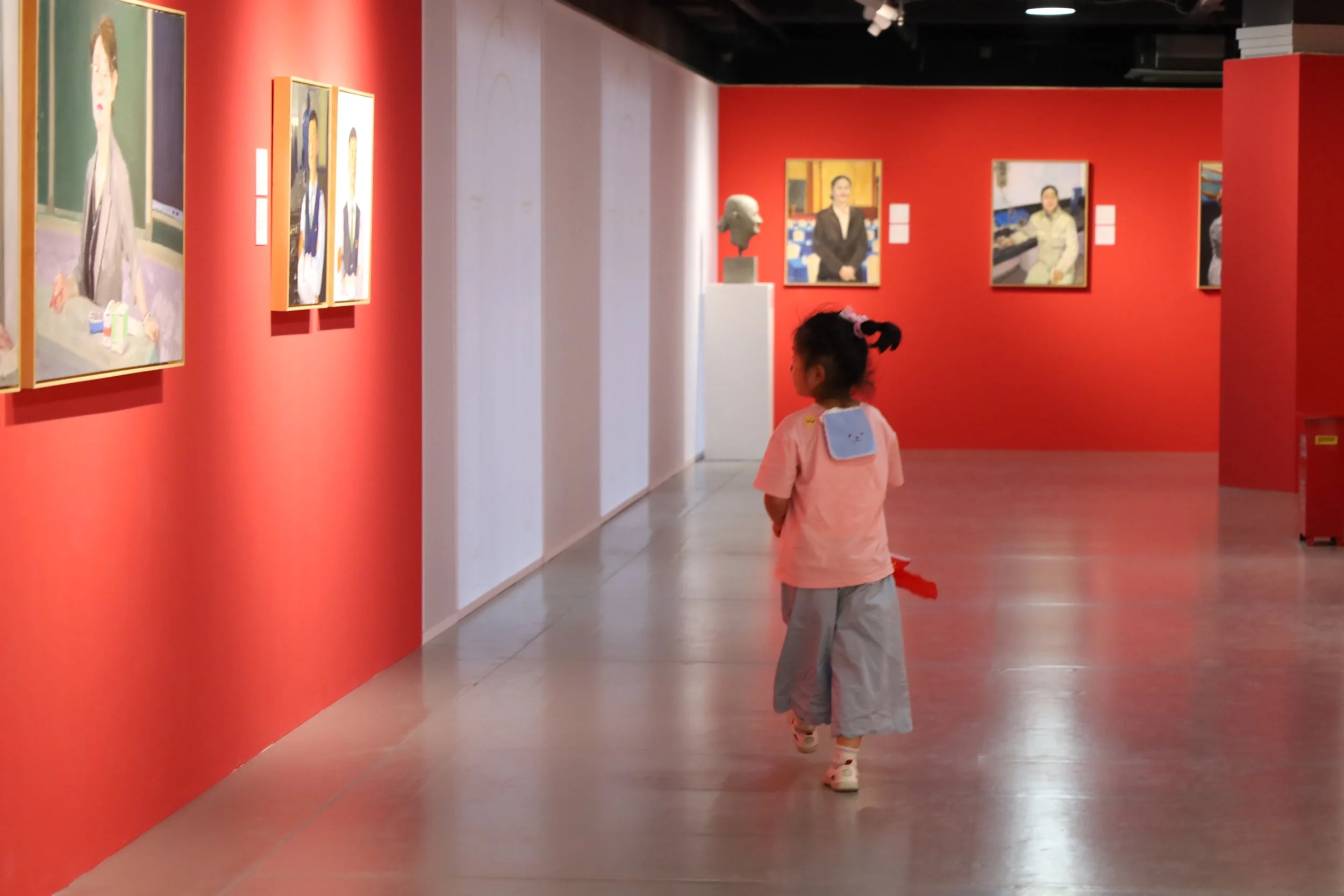 A young girl with a blue backpack walking in an art gallery with red walls and paintings on display.