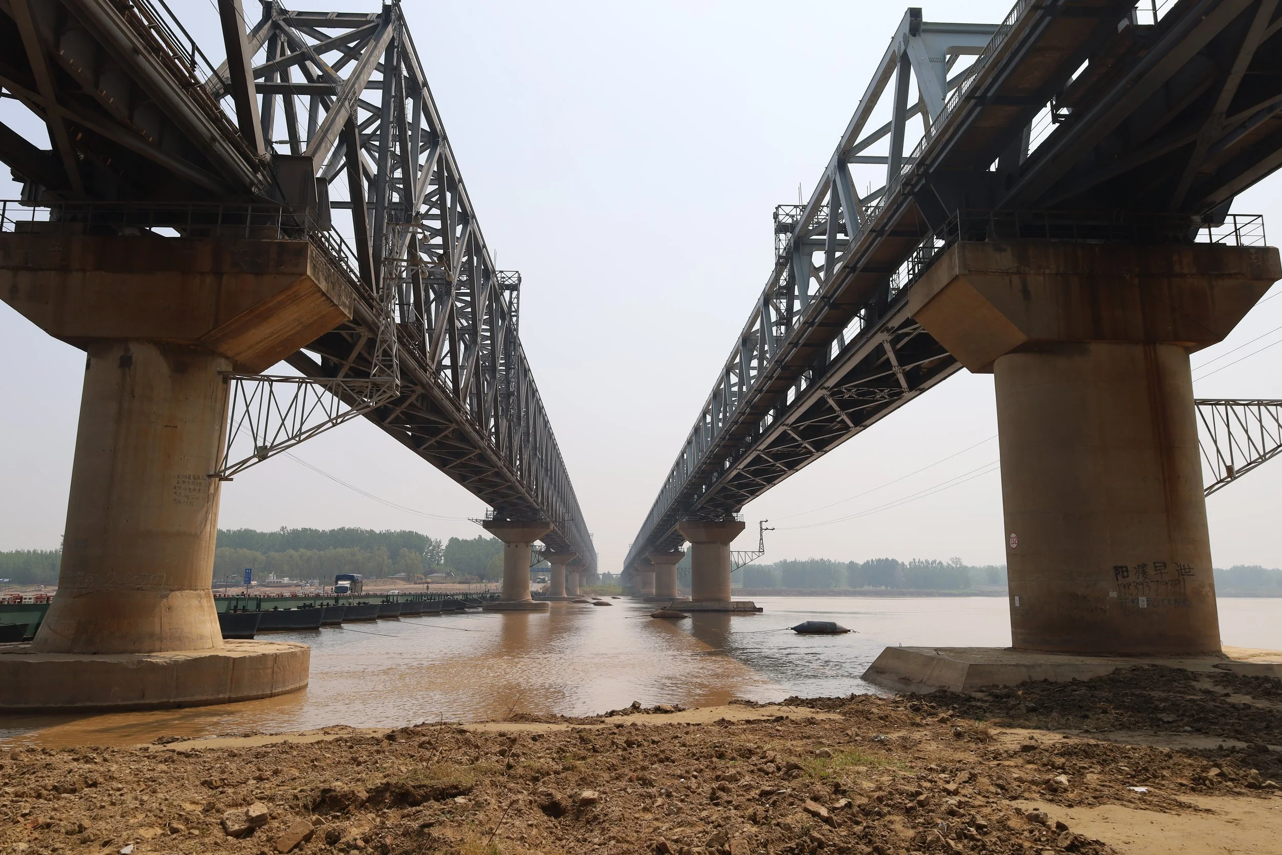 A large steel bridge spans over a body of water, supported by concrete pillars and with an understructure of steel trusses. The sky is overcast, and there is a shoreline with some debris and a few trucks visible in the background.
