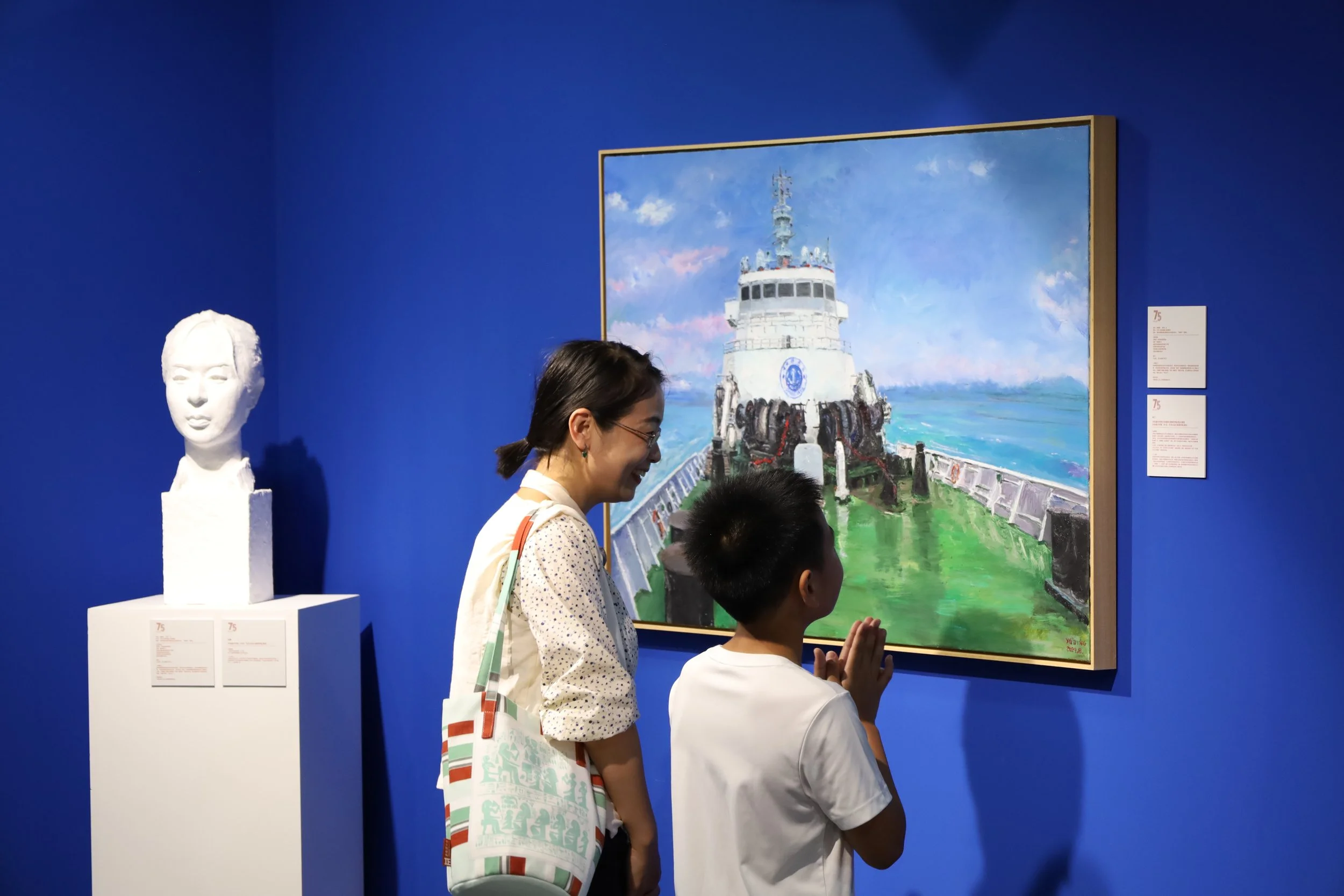 People viewing a painting of a lighthouse at an art gallery with a blue wall background.