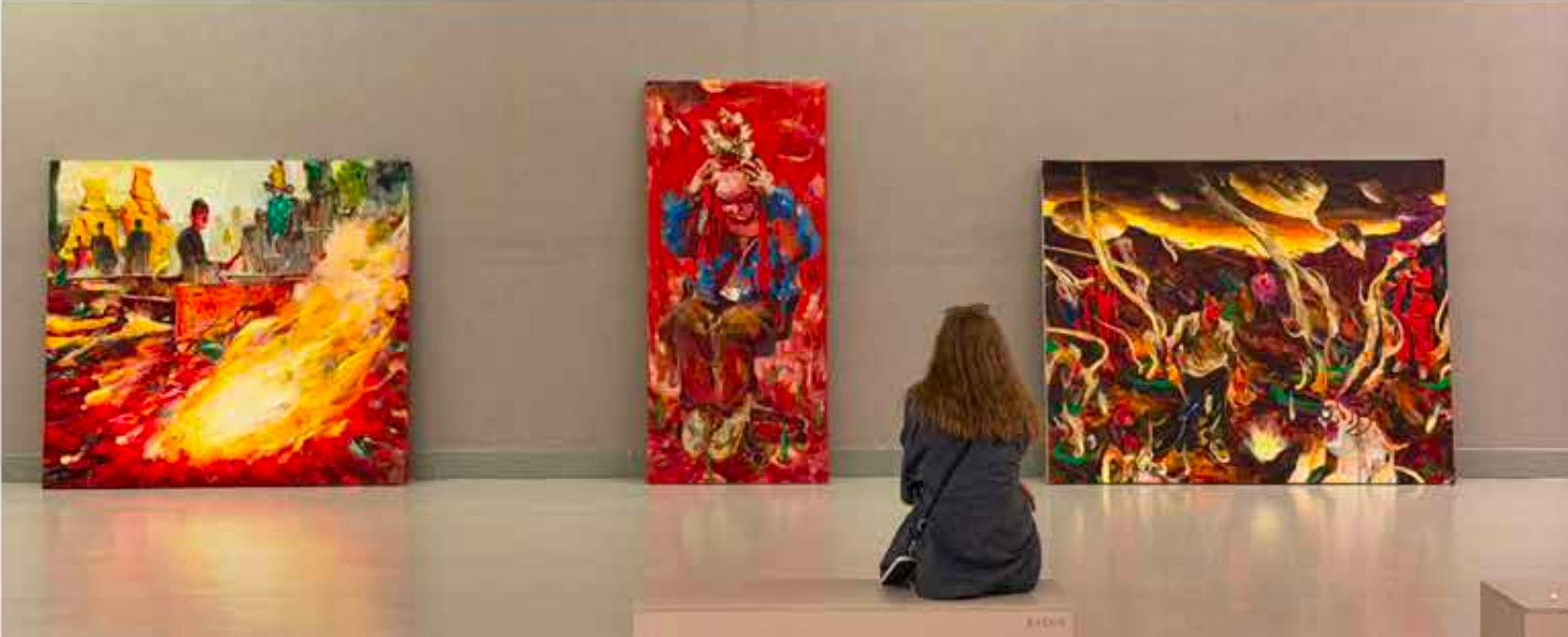 A person with long hair sitting on the floor in front of three colorful paintings in an art gallery.