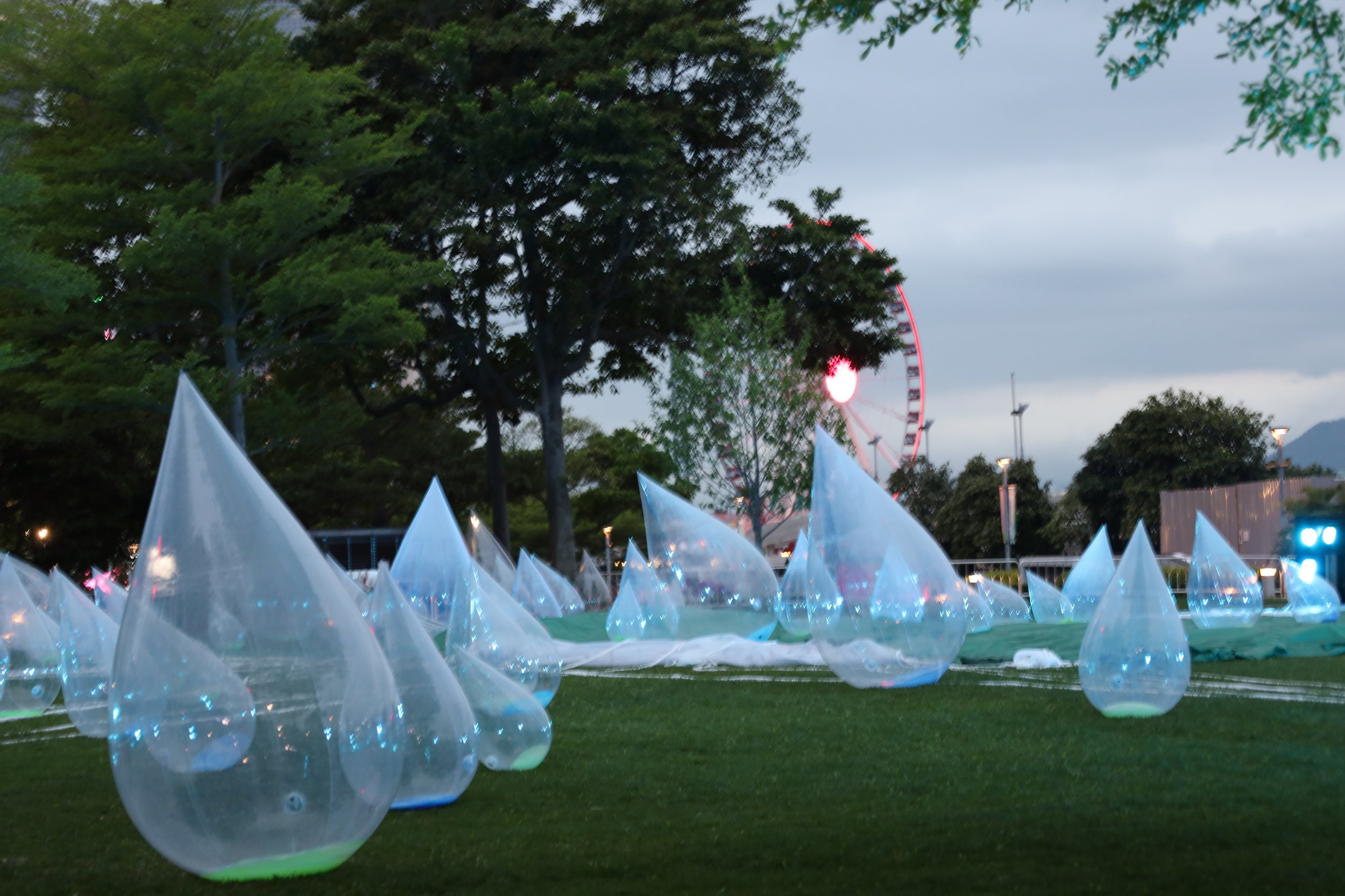View of a ferris wheel behind trees, with illuminated cone-shaped decorations on the ground in a park during dusk.
