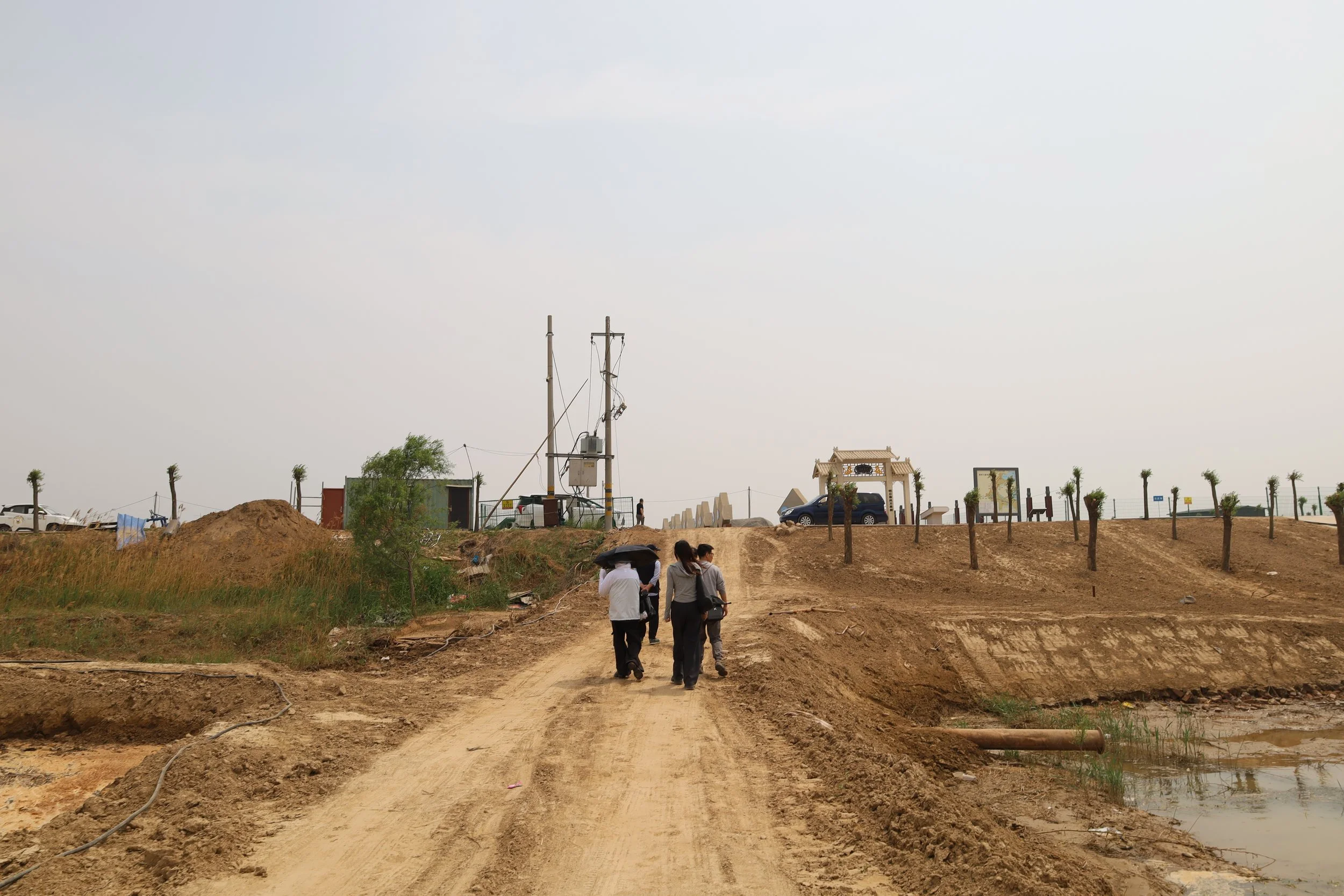 Young artist walking along the road in an underdeveloped area, with sparse trees, utility poles, and a small structure in the background under a cloudy sky.