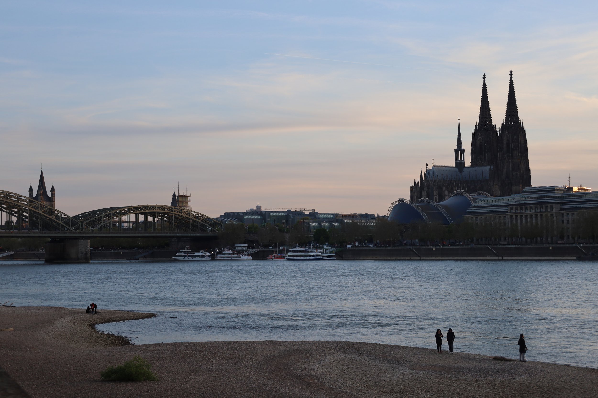 View of the Cologne Cathedral with Seine River in the foreground, featuring a sandy beach, boats, and people walking along the shore during evening.