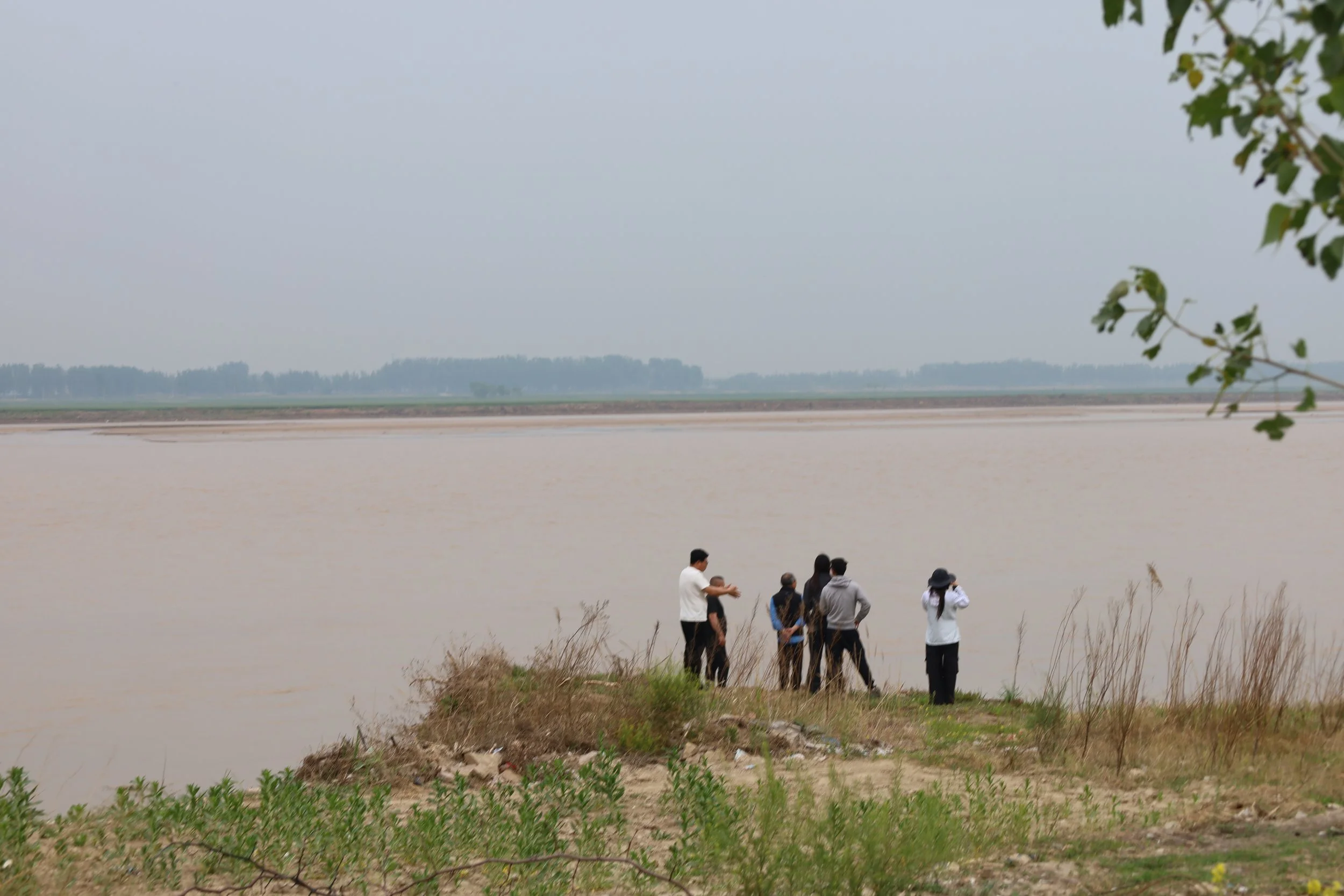 A group of people standing near a body of water, with some taking pictures. The background shows a hazy sky, distant trees, and a flat landscape.