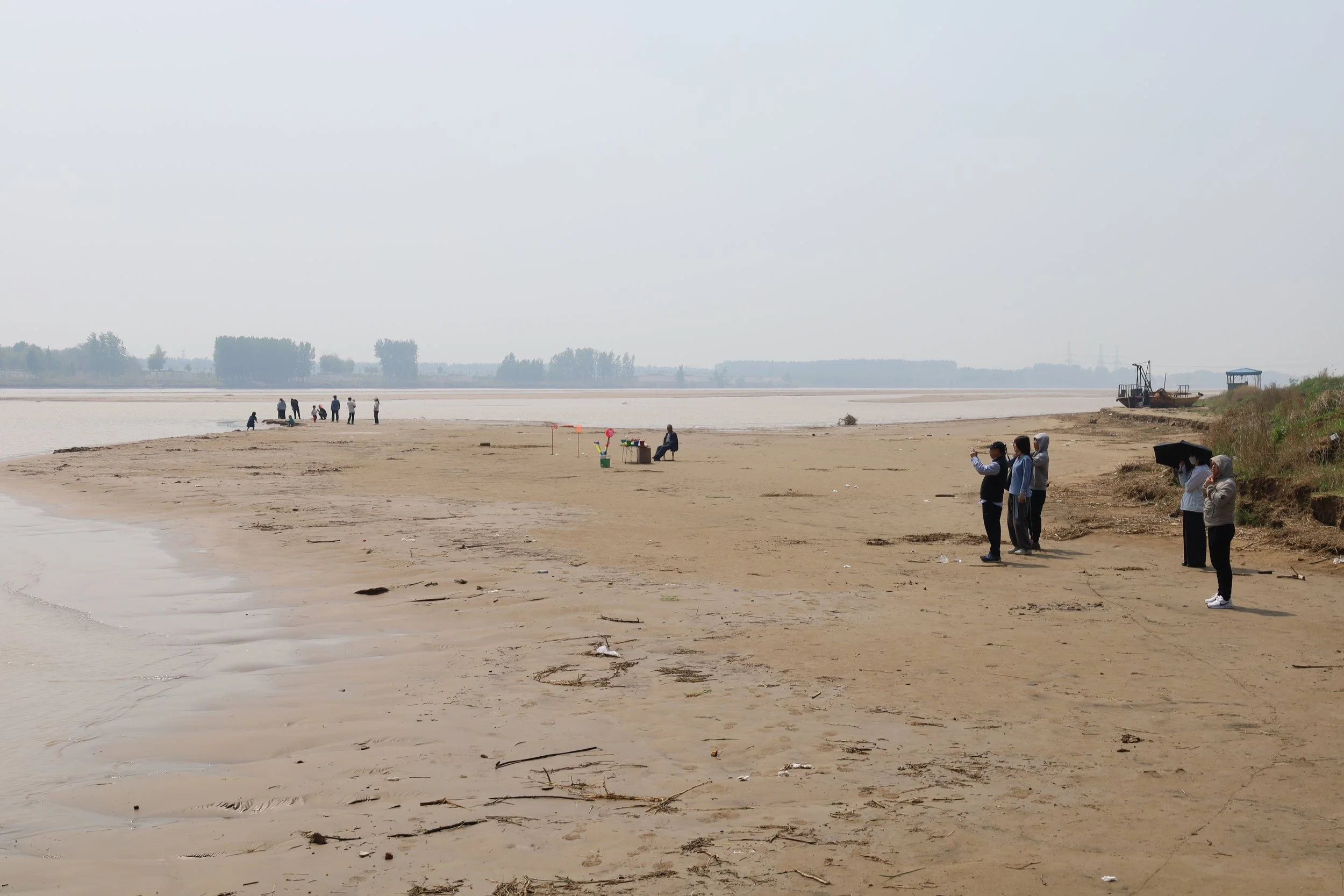 People strolling and sitting along a sandy riverbank or lakeshore on a cloudy day, with trees and a boat in the background.
