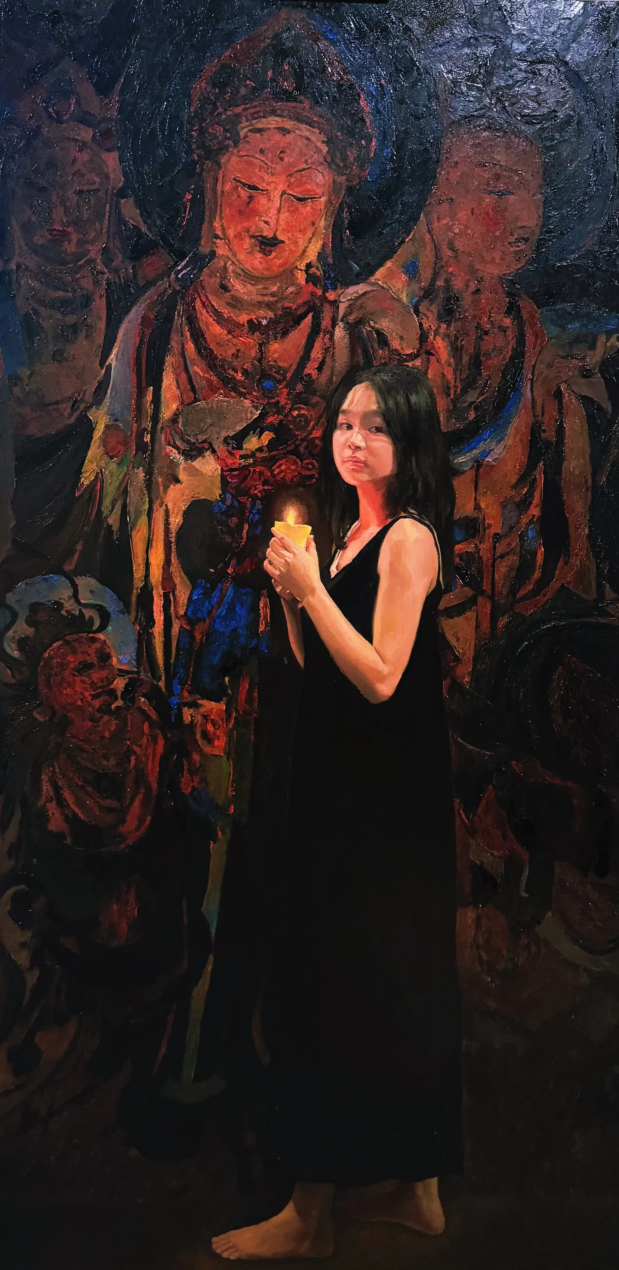 A young woman with long dark hair in a black dress holding a lit candle standing in front of a large mural depicting several faces and skulls.