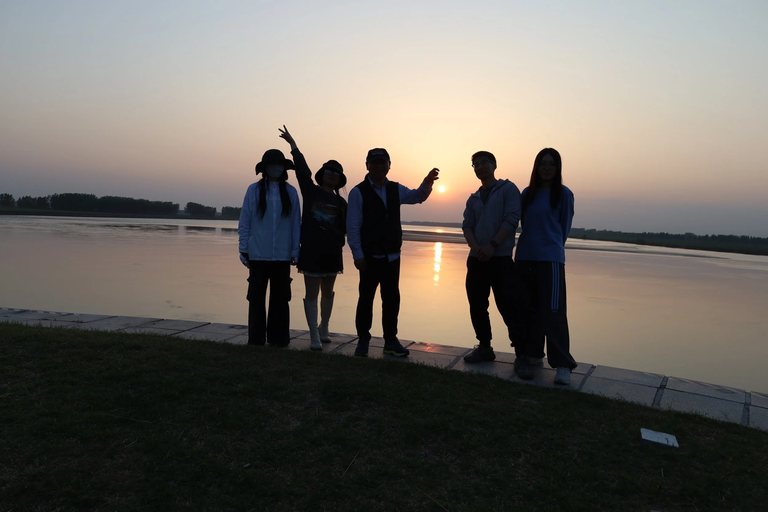 Group of five people standing by a river at sunset, silhouetted against the sky.