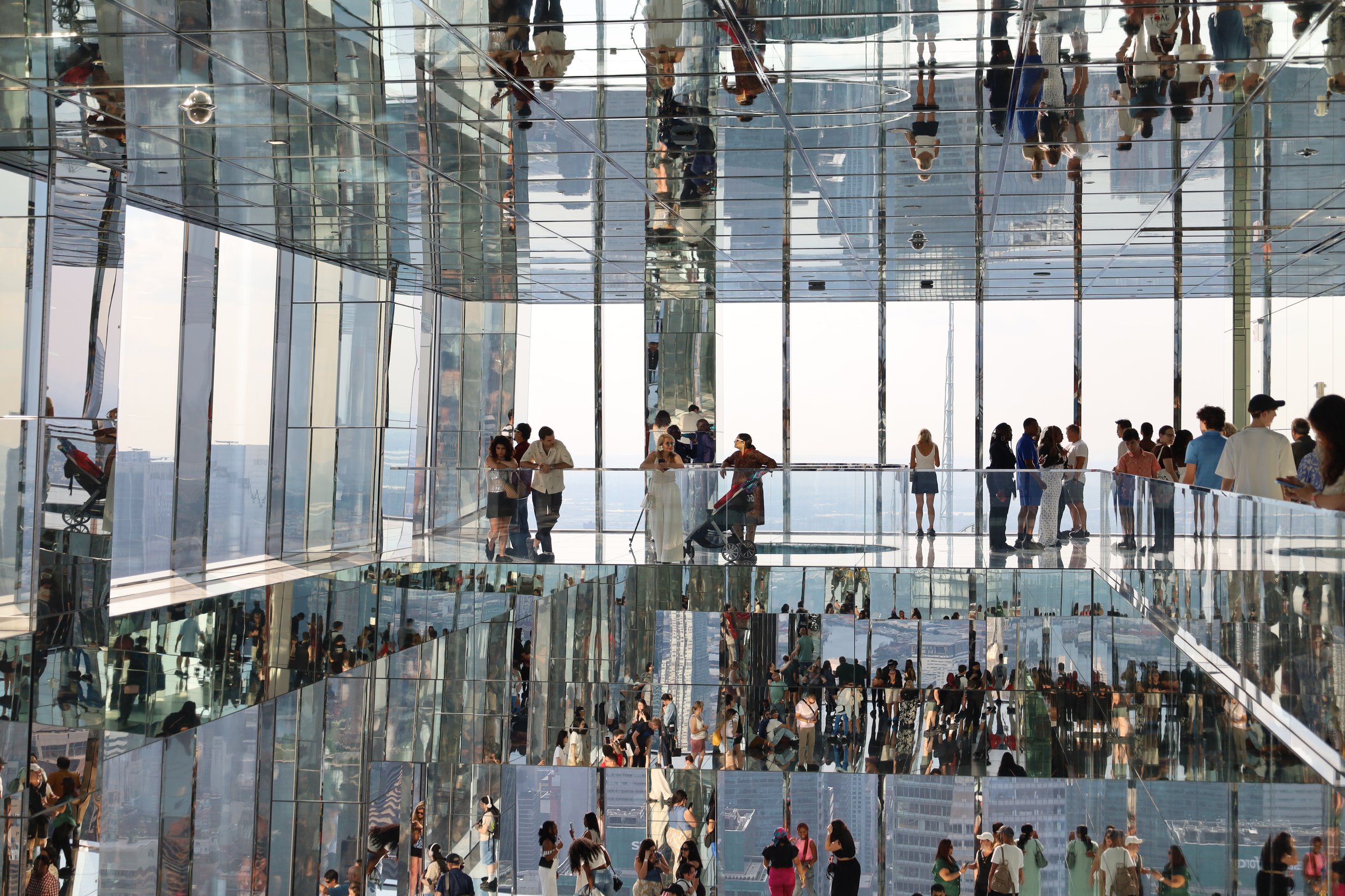 Multiple people walking and standing inside a mirrored, glass-covered building with reflections of the city skyline outside.