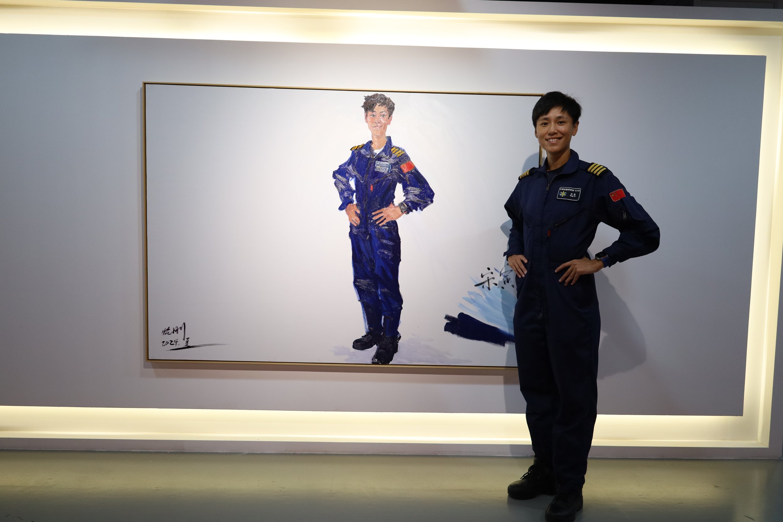 A young woman in a military uniform standing with her hand on her hip, smiling, next to a large framed watercolor portrait of himself in the same uniform, displayed on a wall.