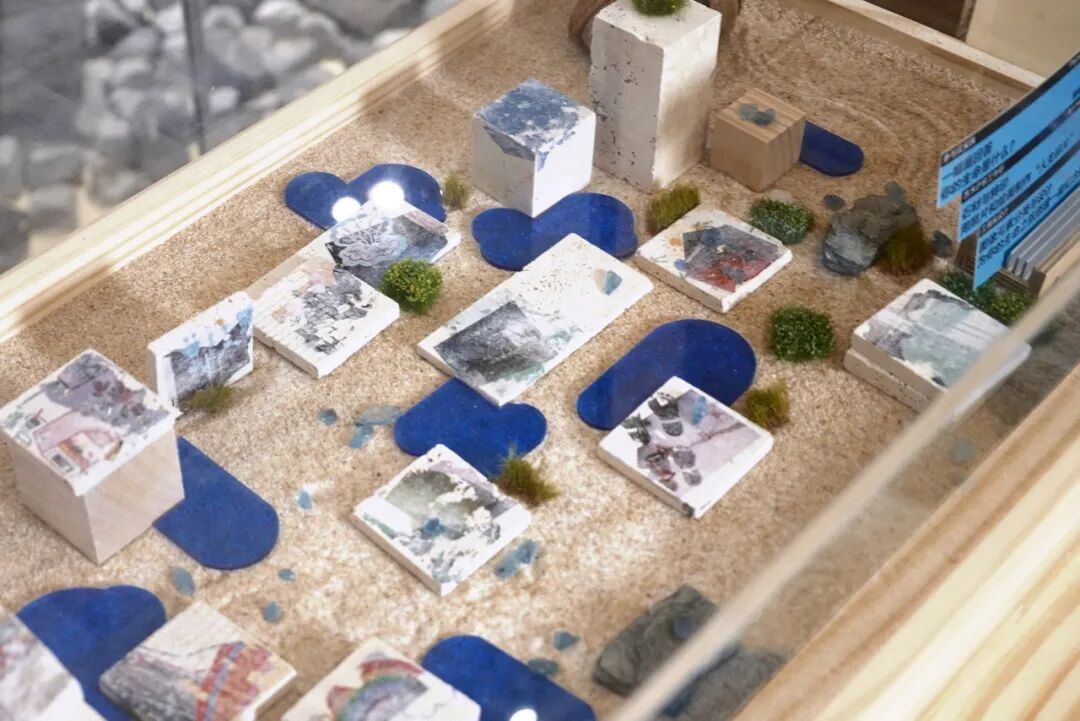 Architectural model of a building complex with white and blue structures, including cubes, rectangular blocks, and circular elements, placed on a sandy ground within a glass display case.