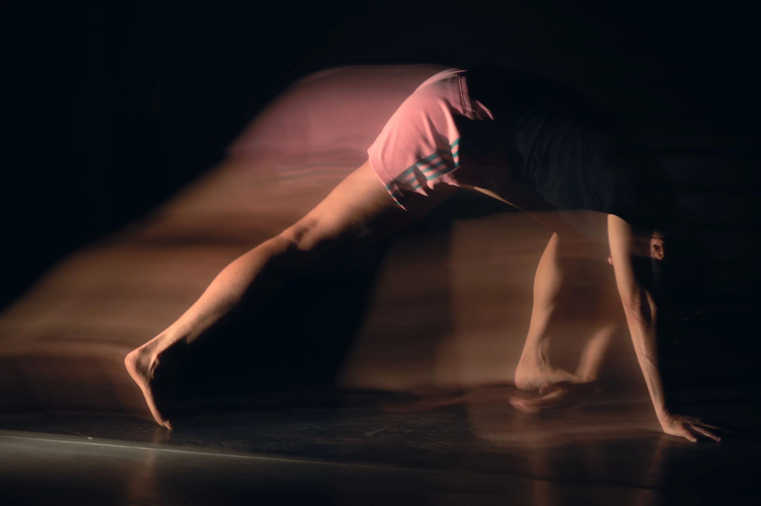 Une personne en mouvement, effectuant une figure de danse dans un espace sombre, portant un t-shirt rose avec des rayures bleues et noires.