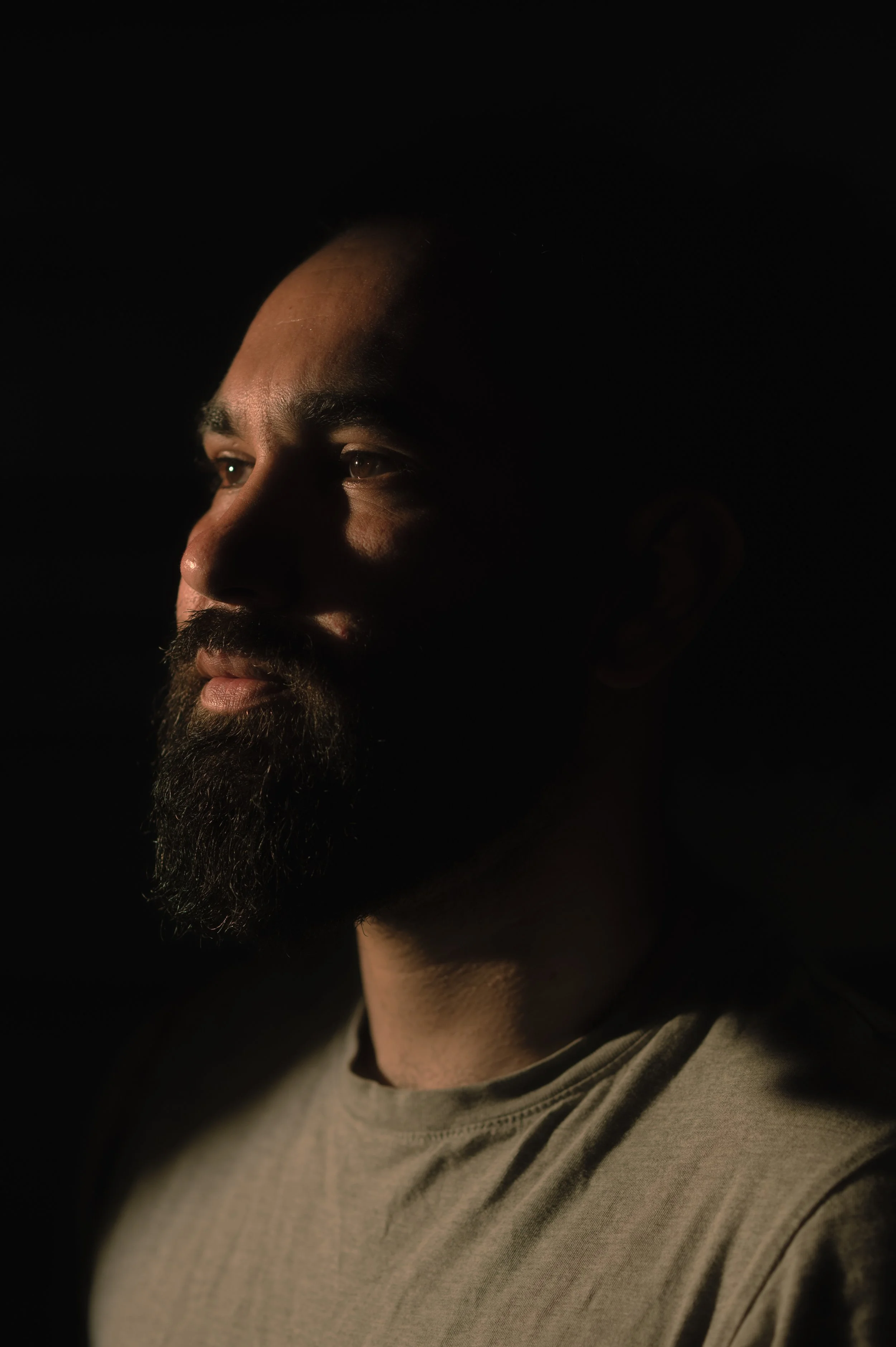 Portrait d'un homme avec une barbe, regardant vers la gauche, vêtu d'un t-shirt gris, éclairé de côté dans un fond noir.
