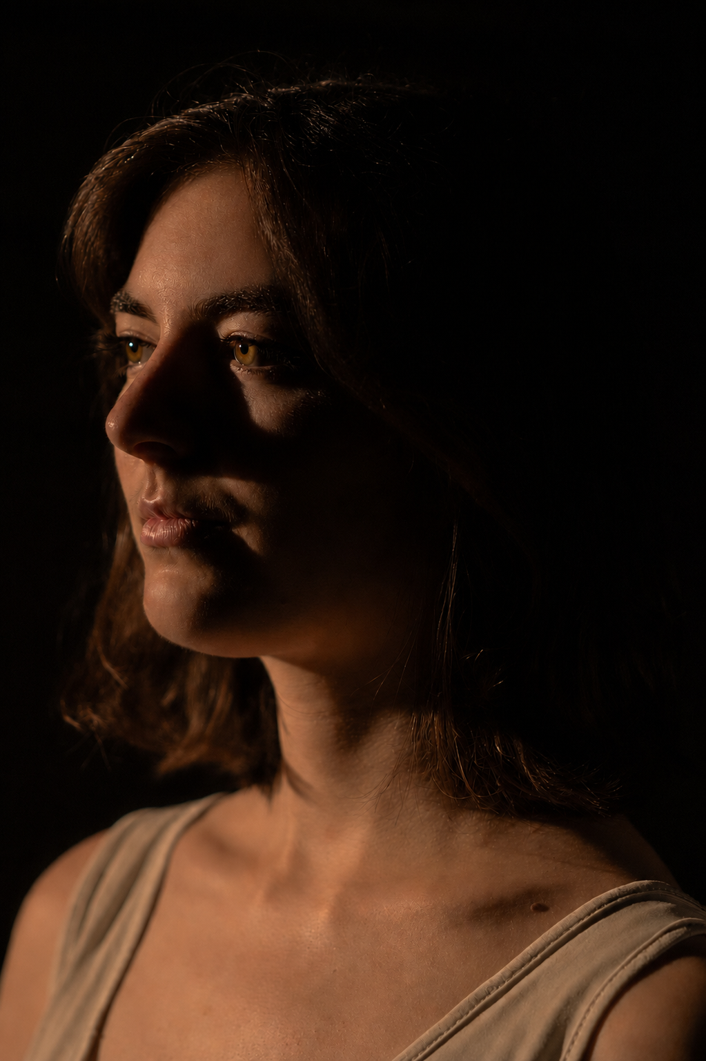 Portrait d'une femme brune, regardant vers la gauche, vêtue d'un débardeur blanc, éclairé de côté dans un fond noir.