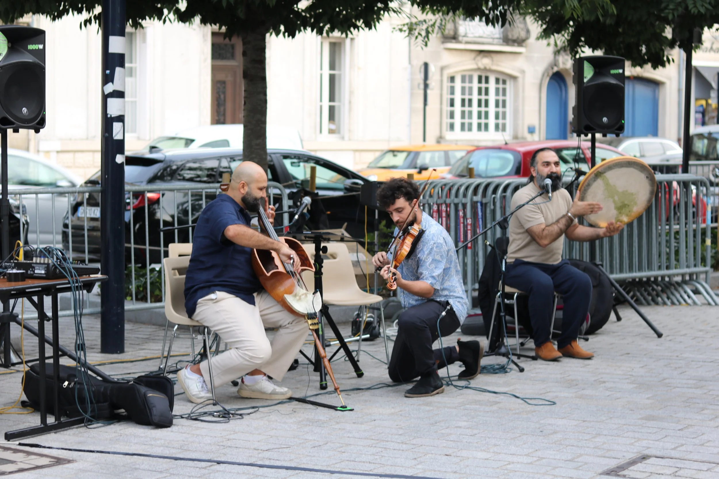 Un groupe de musiciens jouant en plein air dans une place pavée, avec des voitures en arrière-plan. Il y a un homme jouant du geychak, un autre jouant du violon, et un troisième homme avec un tambour. La scène est sous un arbre au centre.