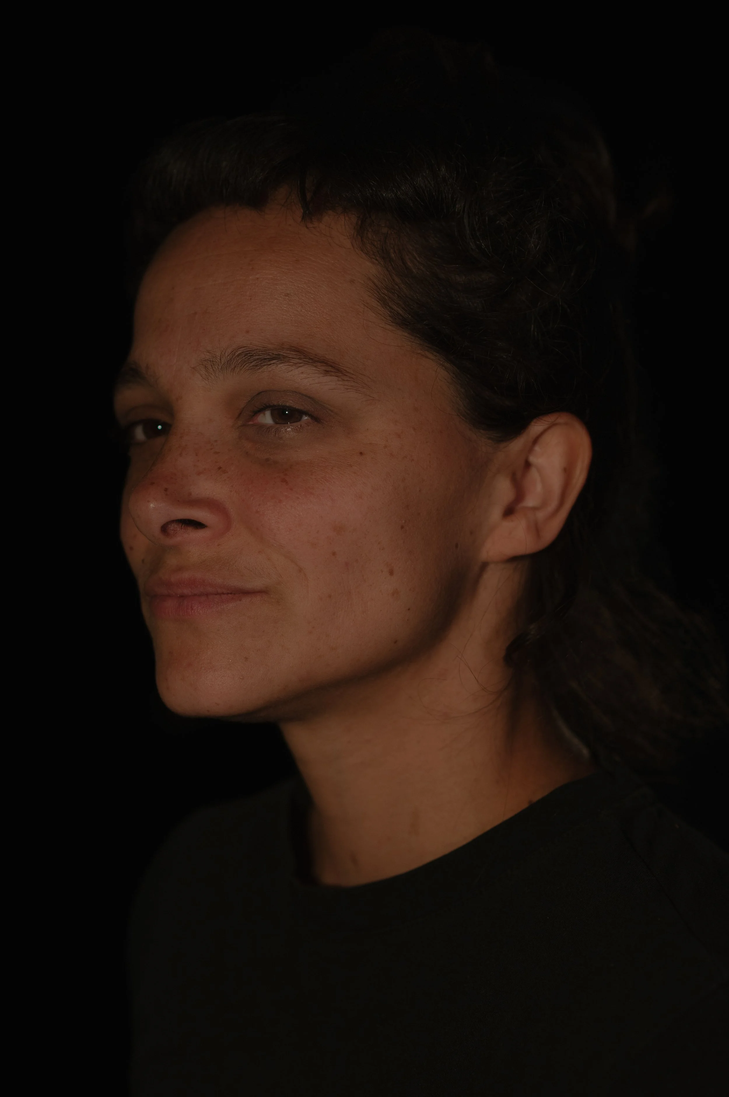 Portrait d'une femme aux cheveux foncés, peau claire avec des taches de rousseur, souriant légèrement, portant un haut noir, fond noir.