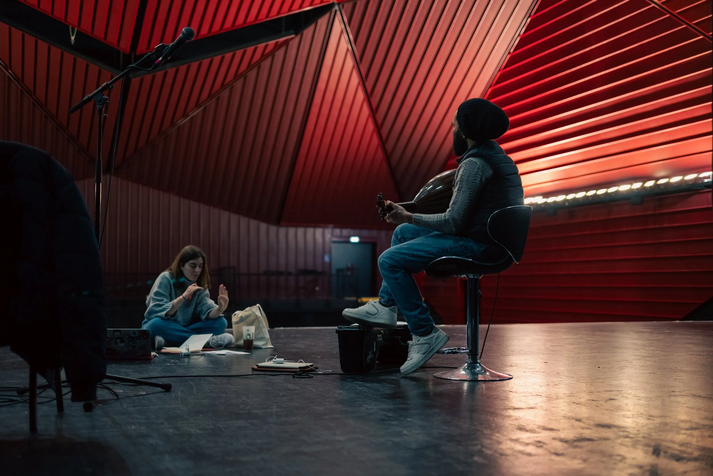 Musicien jouant de l'oud sur scène dans une grande salle au plafond en tôle rouge, Le Rocher de Palmer.