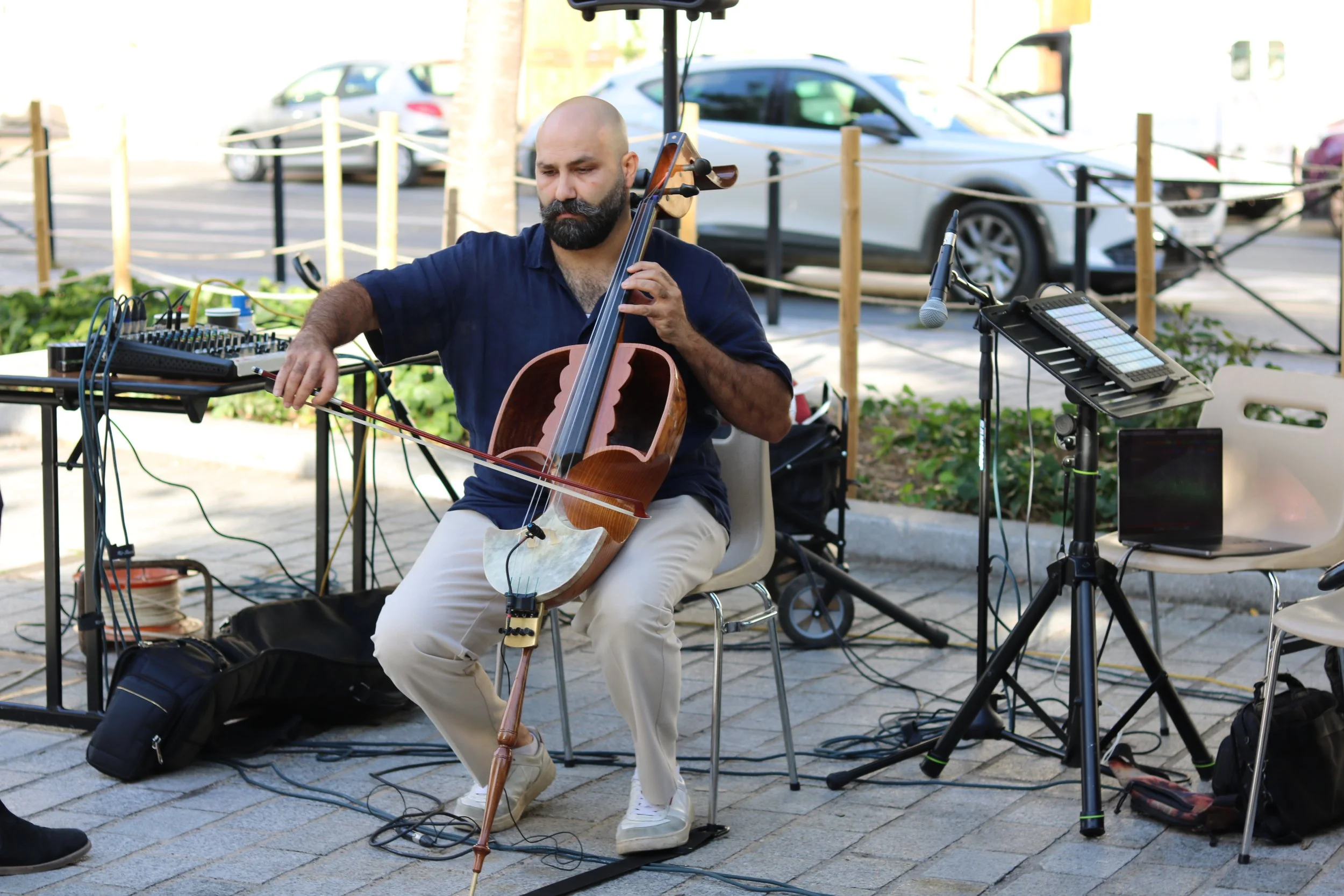 Un homme avec une barbe joue du geychak lors d'une performance en plein air, entouré de matériel musical, dans un espace urbain avec des voitures en arrière-plan.