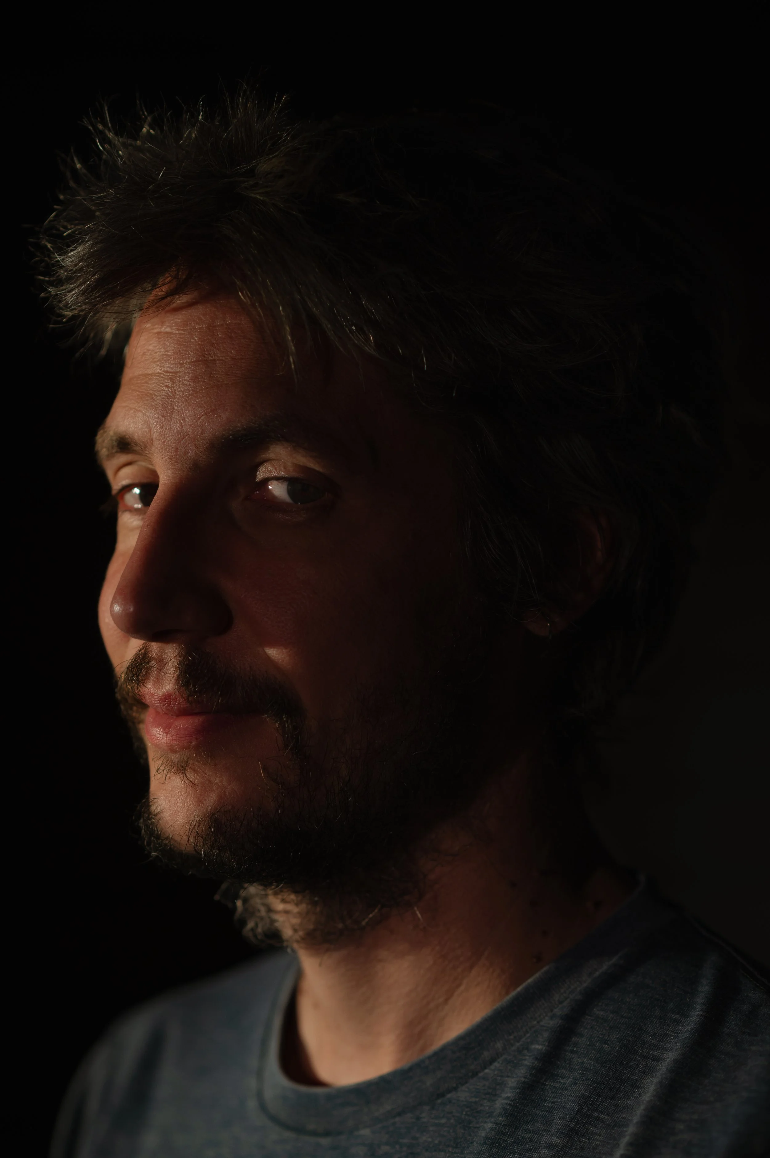 Portrait d'un homme avec des cheveux bruns et une barbe, portant un t-shirt gris foncé, regardant la caméra et souriant légérement, éclairé de côté dans un fond noir.
