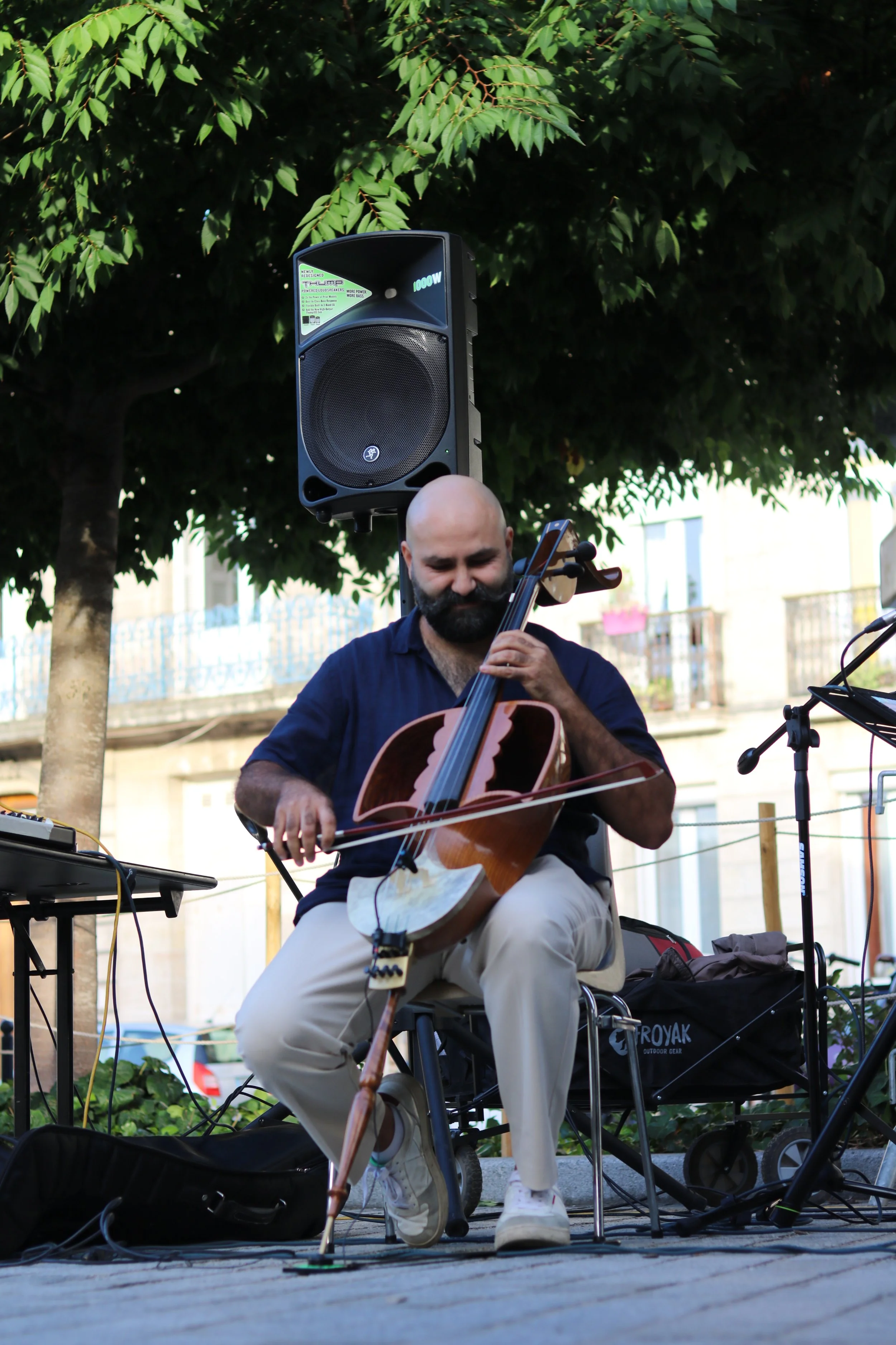 Un homme joue du geychak (instrument rare à cordes) lors d'une performance en plein air, sous un arbre, avec un équipement audio et électronique visible autour de lui.