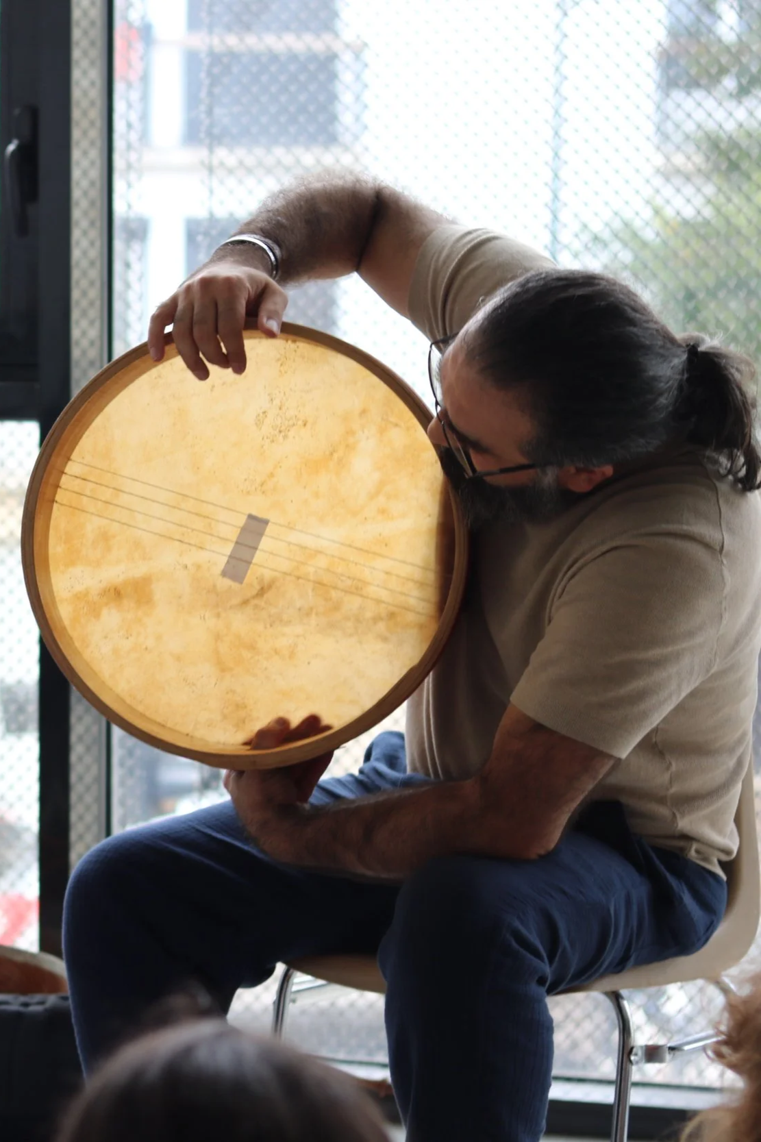 Homme montrant un daf (grand tambourin), portant des lunettes et une chemise beige, dans une pièce avec une fenêtre en arrière-plan.