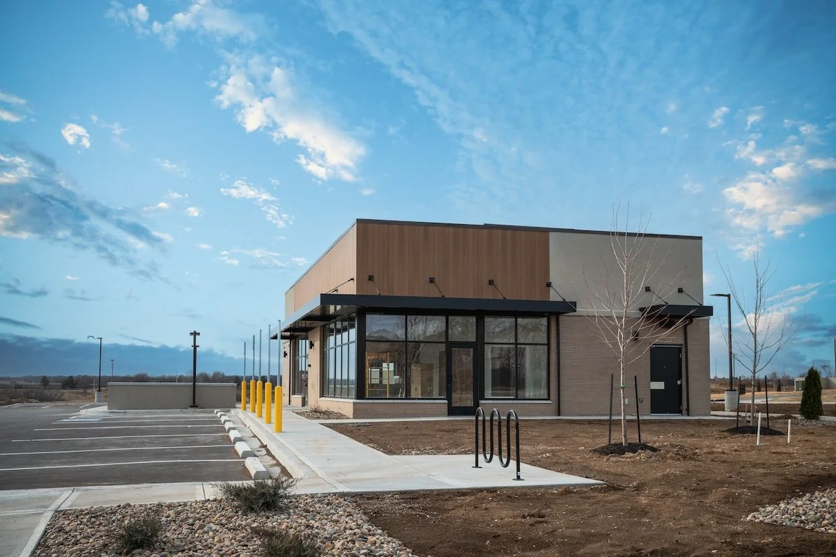 New commercial building with large glass windows, parking lot, and a few leafless trees in front, under a partly cloudy sky.
