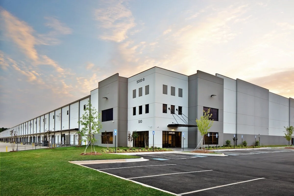 Large white modern warehouse or industrial building with neatly landscaped parking lot and few small trees, under a partly cloudy sky at dusk or dawn.