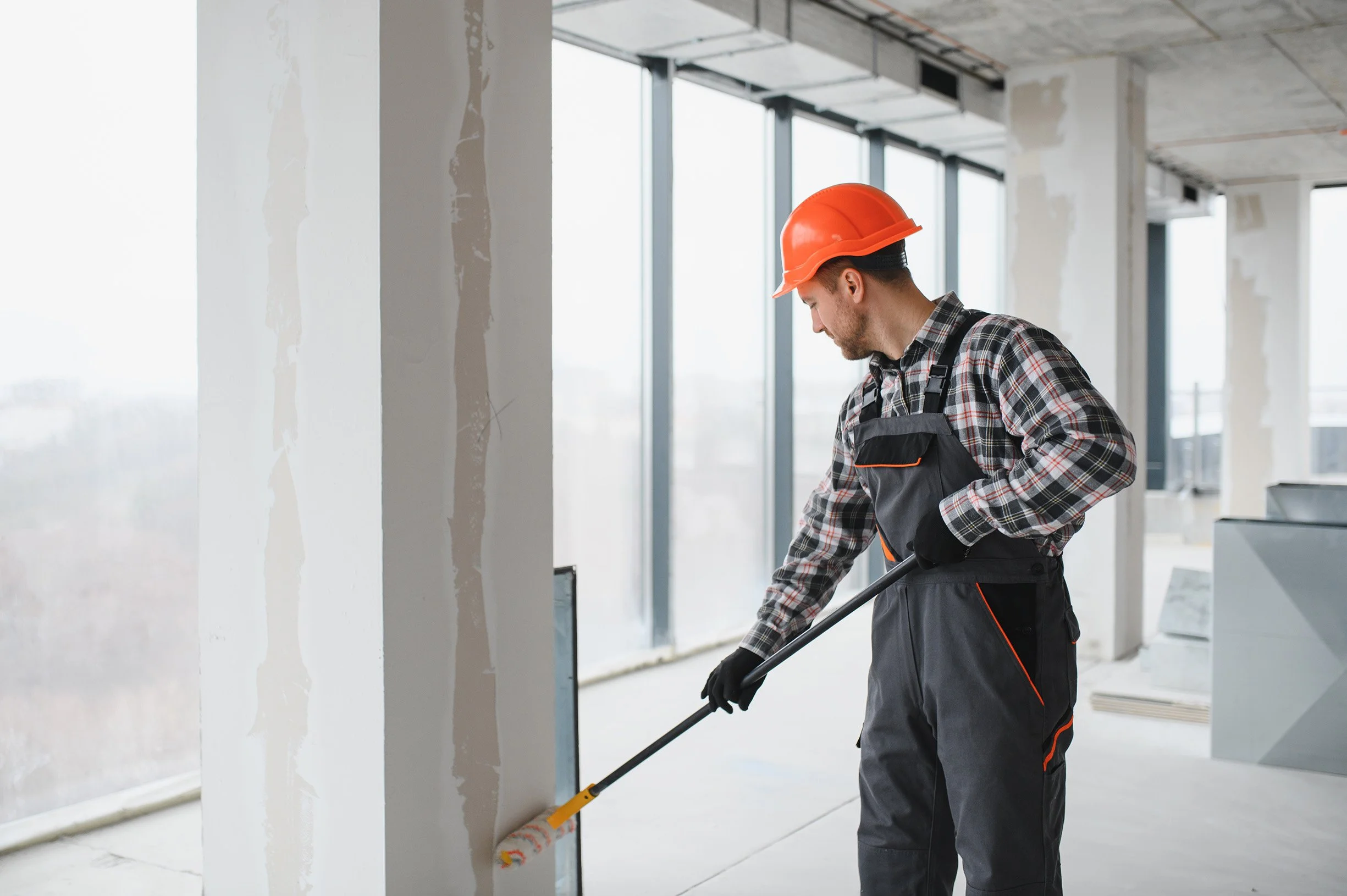 Construction worker in a plaid shirt, black overalls, orange safety helmet, and gloves sweeping a smooth concrete floor inside a building with large windows and partially finished walls.