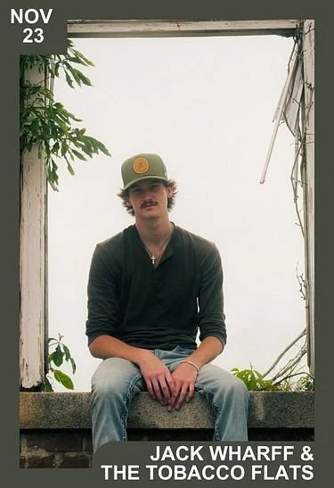 Young man with a mustache and beard, wearing a green baseball cap, black long-sleeve shirt, jeans, and a necklace, sitting on a concrete ledge in front of a broken window with plants around him.
