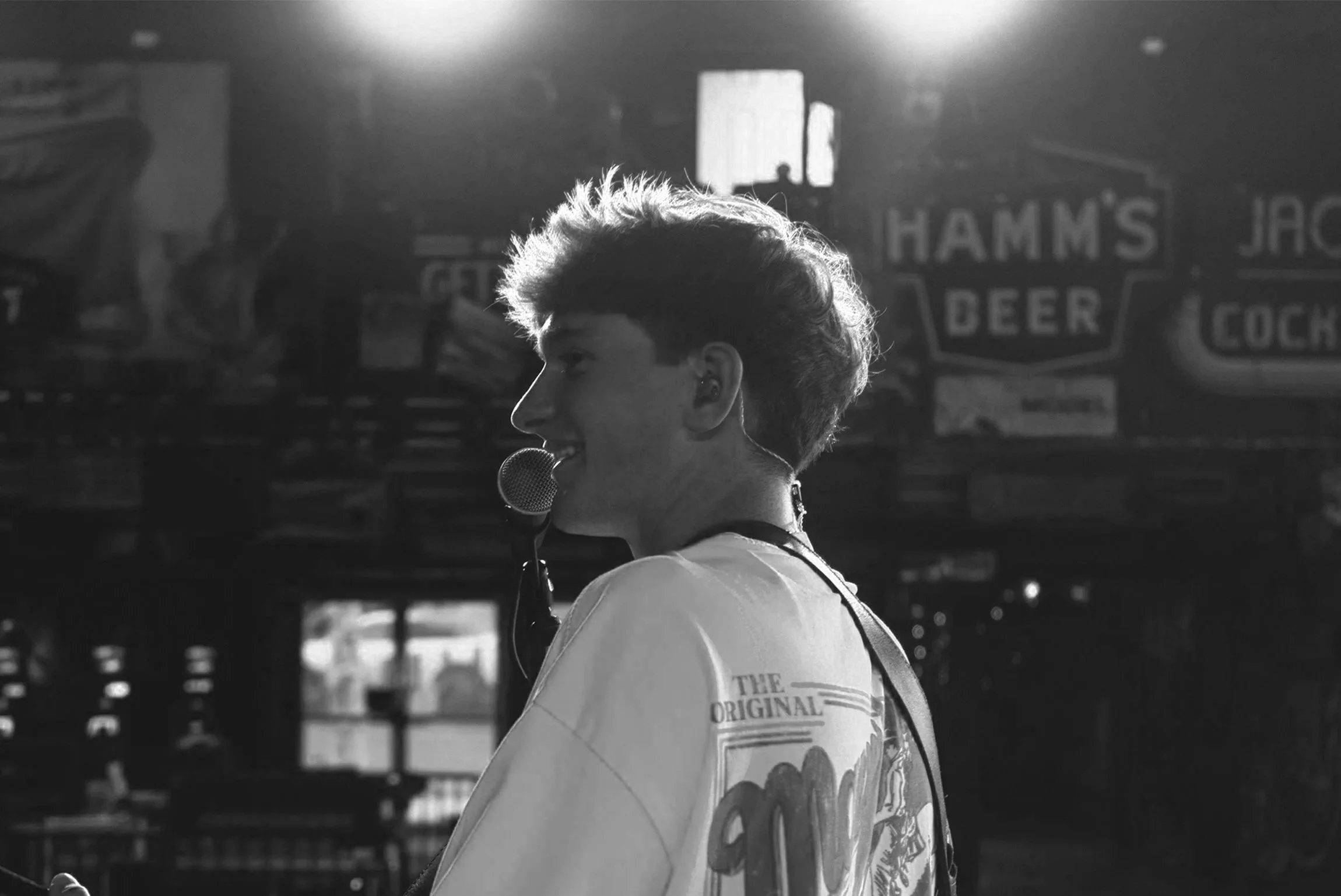 A black and white image of a young person with short, curly hair wearing a t-shirt with a graphic design, talking into a microphone. The background includes signs for a bar and other shops, with sunlight streaming through windows.