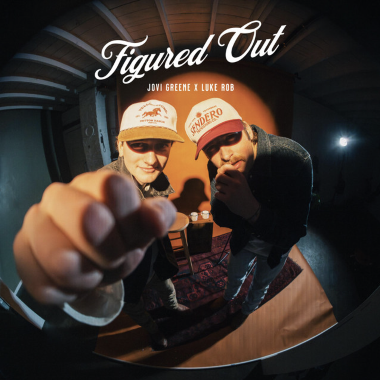 Two men wearing caps and sunglasses look into a fisheye camera while standing in front of a sign that says 'Figured Out' with the names 'Jovi Greene x Luke Rob'. One man reaches towards the camera and the other is touching his lips, with a table and small objects behind them.