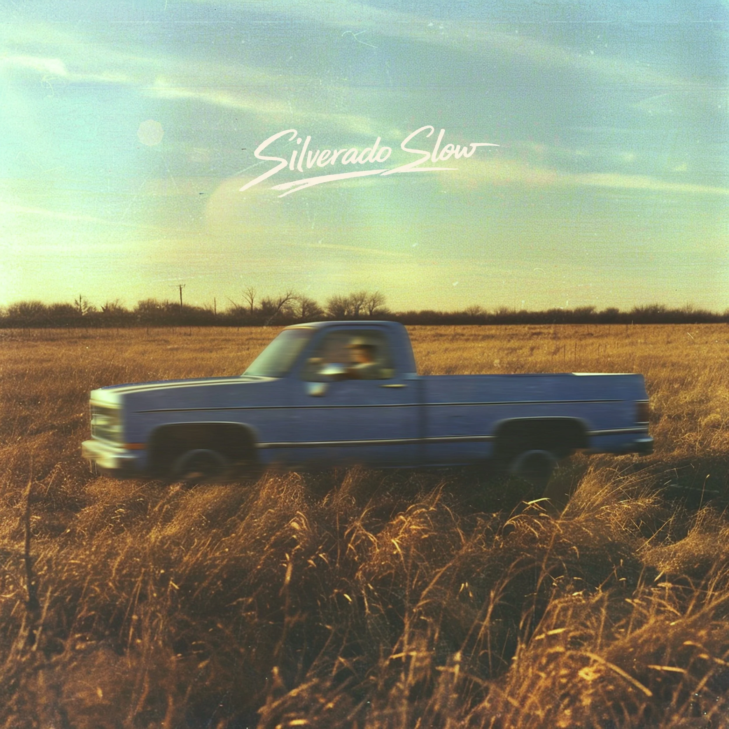 A blurry photo of a moving vehicle with a landscape of open fields and trees. The sky is blue with some wispy clouds. The photo has a bright, slightly washed-out appearance with visible lens flare.
