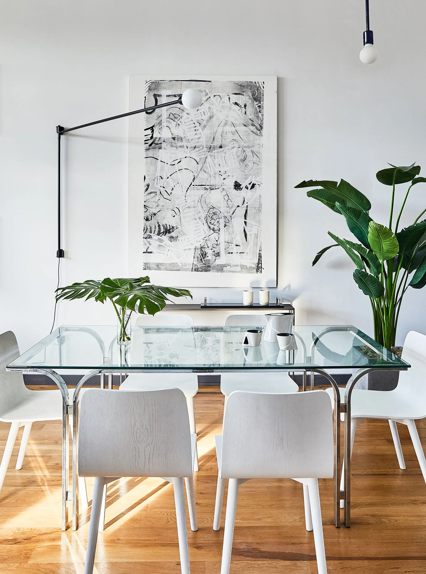 Modern dining room with a glass-top table and six white chairs. Large abstract black and white artwork on the white wall. Potted plants with large green leaves on either side of the table. Minimalist décor with three candles on a sideboard and an exposed bulb hanging from the ceiling.