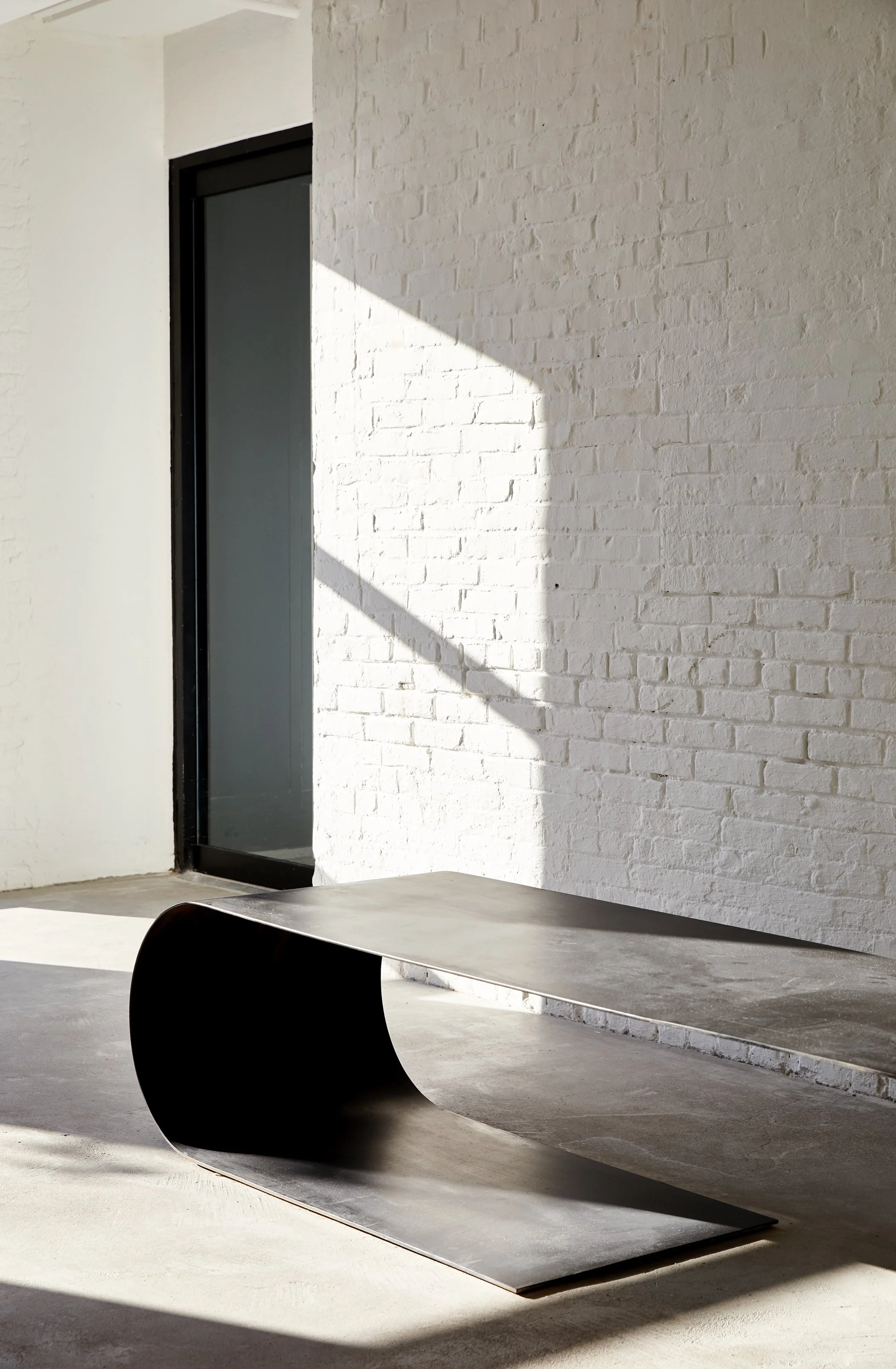 Modern minimalist interior with white brick wall, black elevator door, black curved bench, and concrete floor.