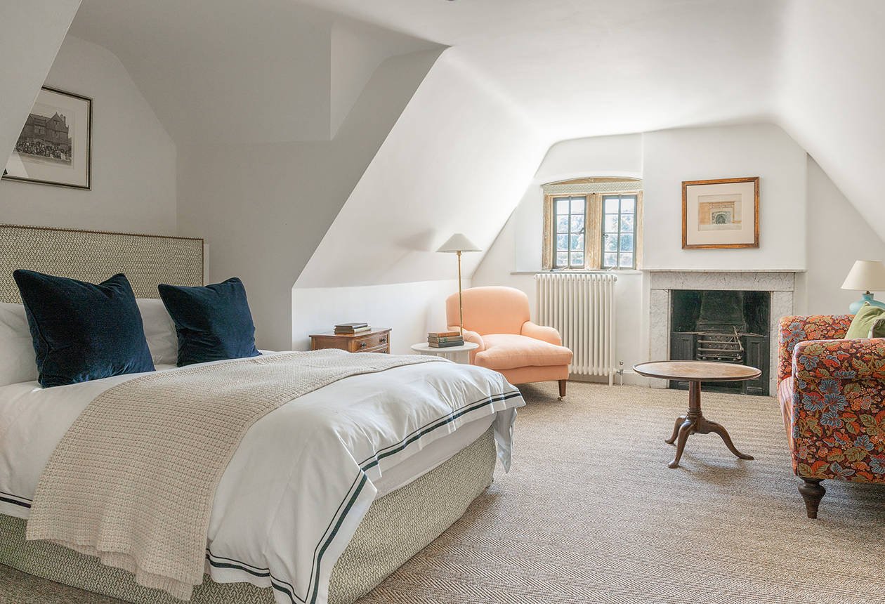 A cozy bedroom with a large bed, navy pillows, and a textured beige headboard. There is a small wooden nightstand with books and a white table lamp. Next to the window, a peach-colored armchair with a floor lamp beside it. In front of the window, a fireplace with a framed artwork above it. To the right, there is a floral-patterned armchair with a green pillow and another lamp. A round wooden coffee table is in the center of the room, all on a beige striped carpet.