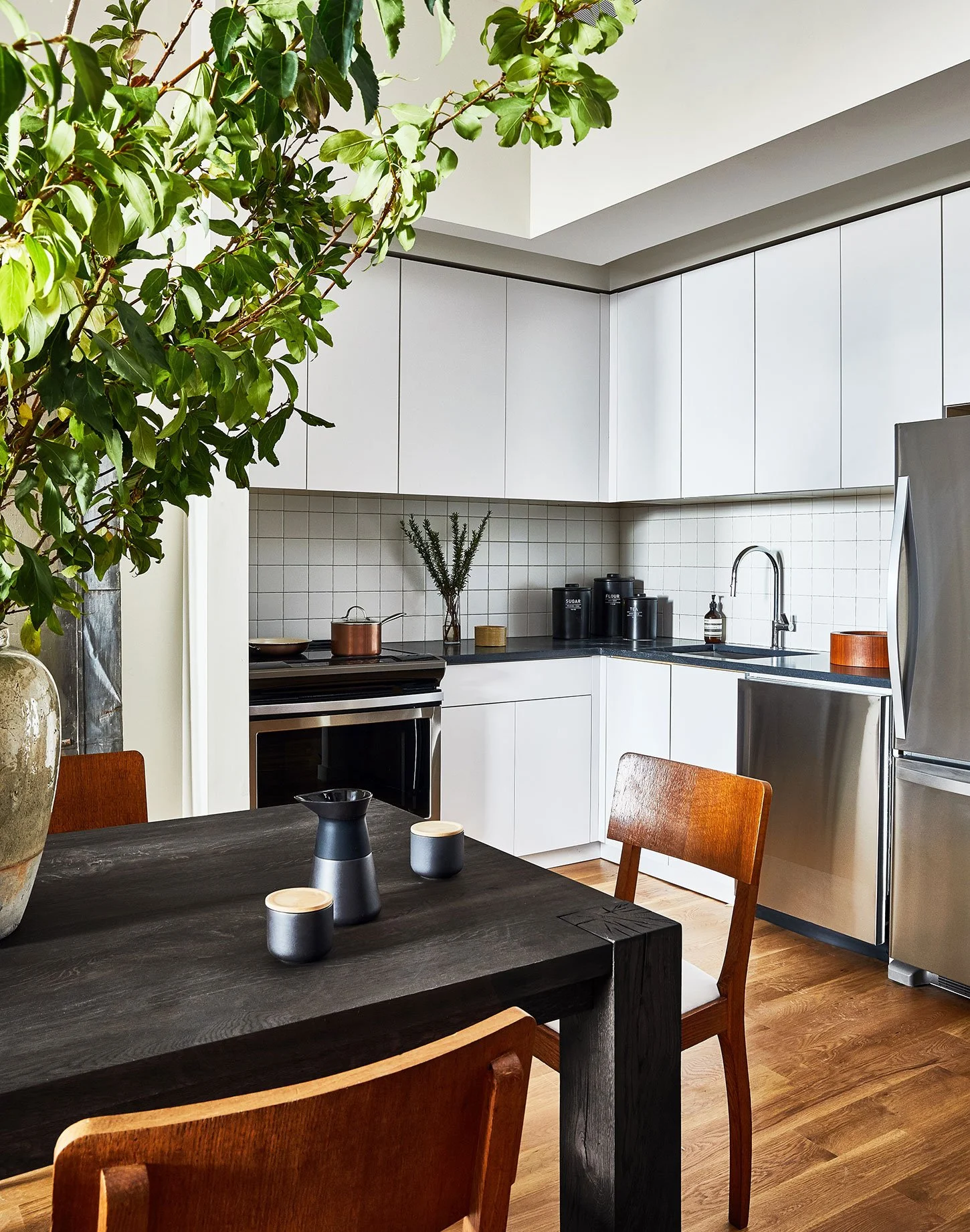 A modern kitchen with white cabinets, black countertops, a stainless steel refrigerator, a black stove with copper pots, and a black tile backsplash. There is a large green leafy plant in a rustic vase on the dining table, which is dark wood with matching chairs. Decorative black and beige containers and a small glass bottle are on the counter near the sink.