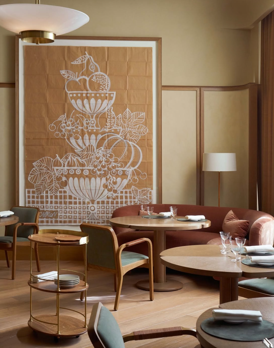 A cozy restaurant interior with tables, chairs, and a large artwork featuring white line drawings of fruit and leaves on an orange background.