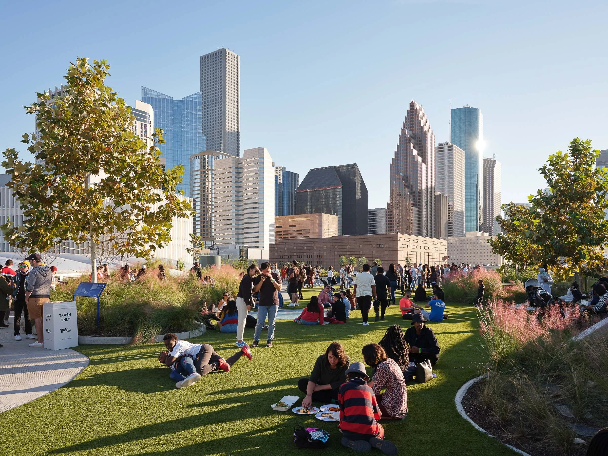 People gathering in a city park with tall skyscrapers in the background on a sunny day, some sitting on the grass and others walking.