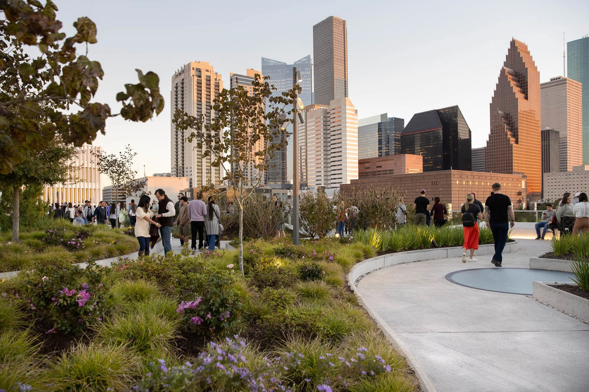 A city park with people walking and socializing, with tall skyscrapers in the background during sunset.