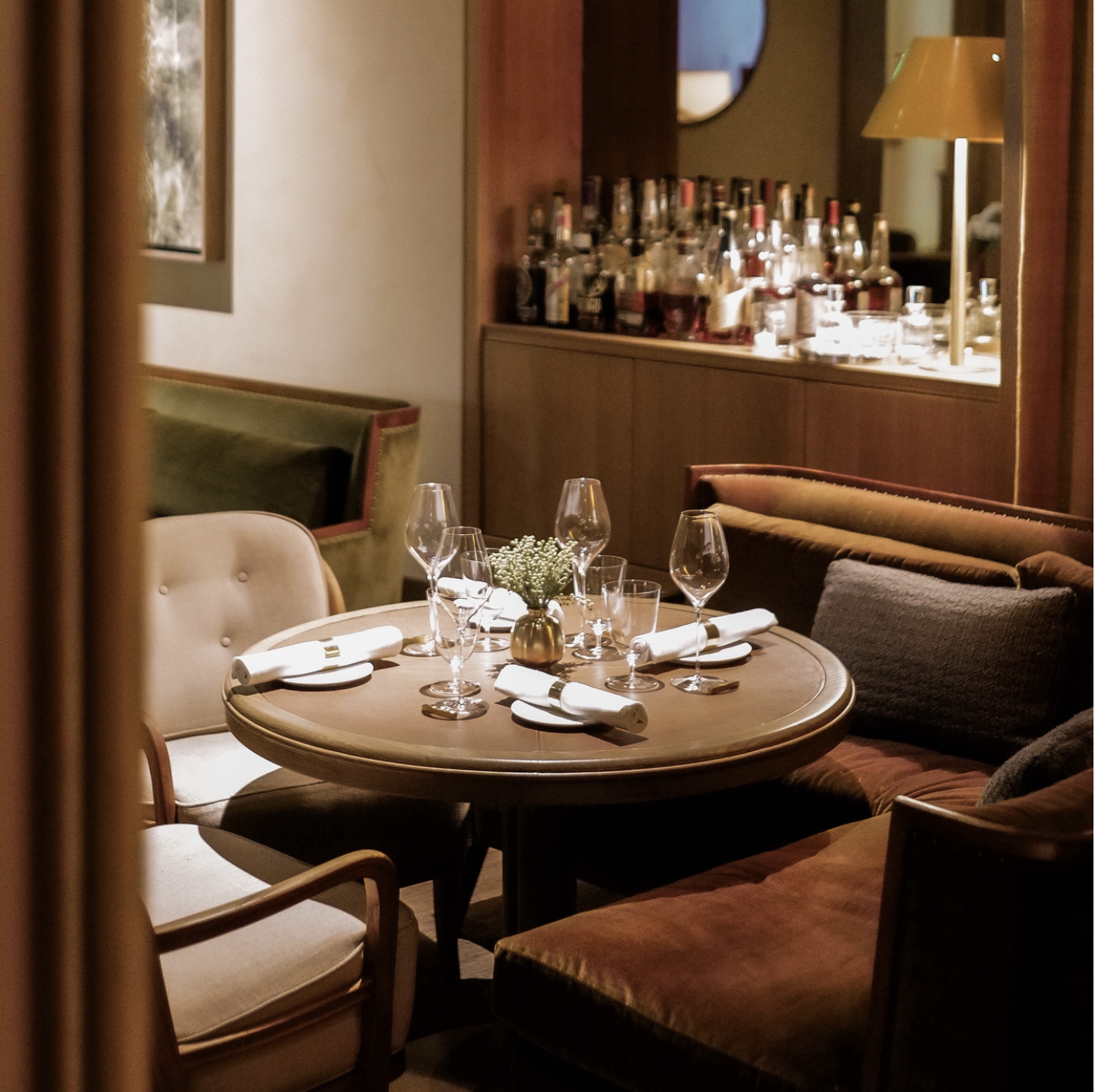 A cozy restaurant table set with wine glasses, white napkins, cutlery, and a small flower in a brass vase, with a bar counter and soft seating in the background.
