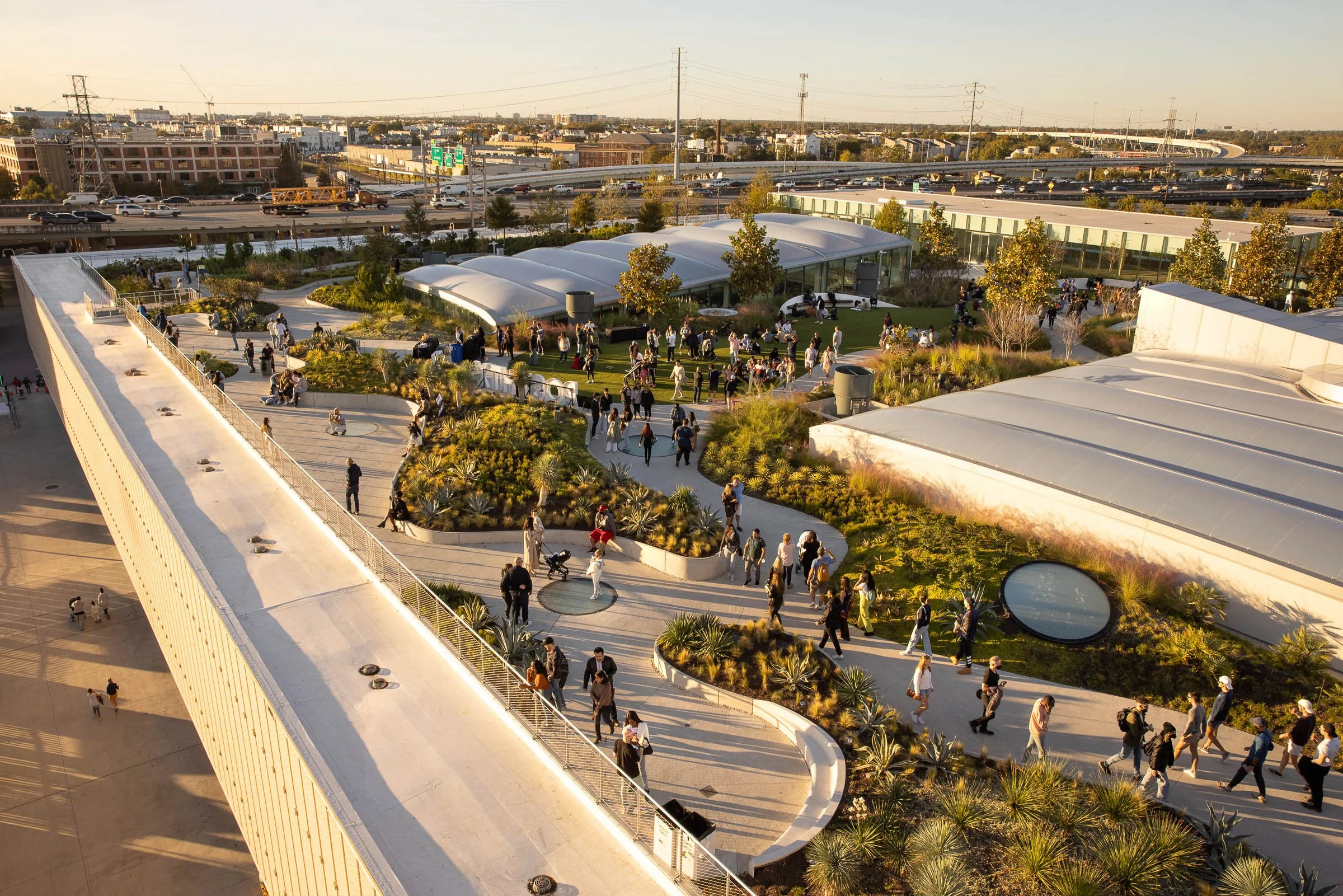 An aerial view of a modern outdoor urban park with many people walking, sitting, and socializing among landscaped gardens, pathways, and glass structures, with highways and city buildings in the background.