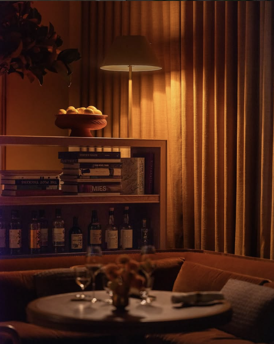 A dimly lit dining room with a round table set with wine glasses and napkins, a cozy sofa, a wooden bookshelf filled with books and liquor bottles, a bowl of lemons on top of the bookshelf, a table lamp casting warm light, and gold curtains in the background.